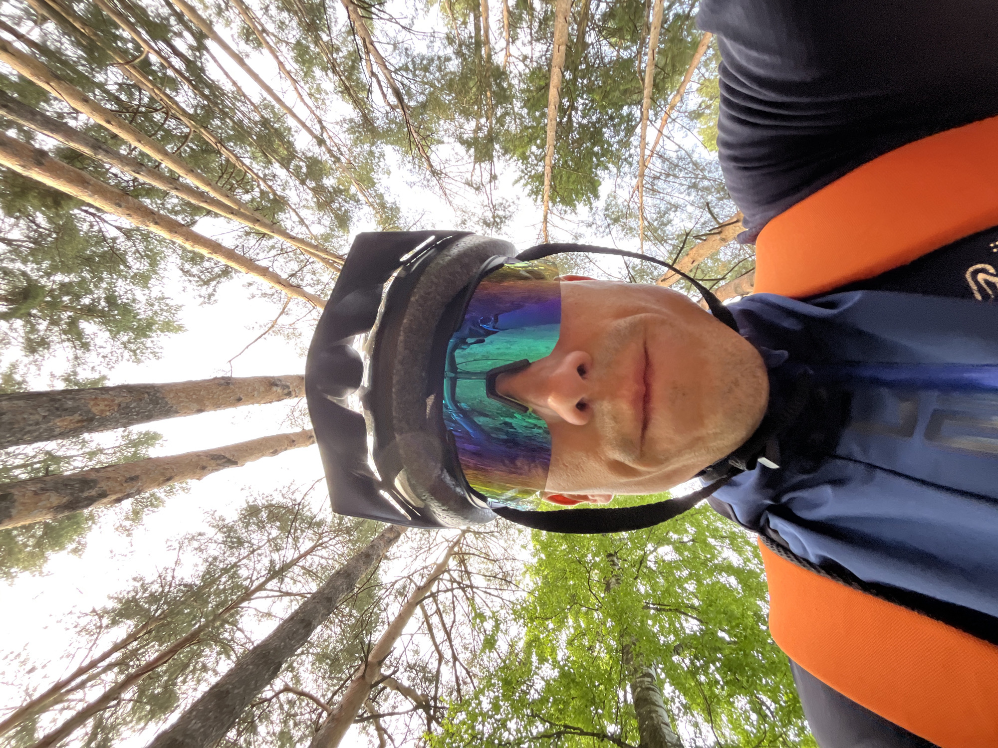 A low-angle selfie perspective captures a person wearing a black cycling helmet and iridescent sunglasses, positioned against a backdrop of tall trees. The person is also wearing a blue cycling jersey with an orange detail on the upper shoulder/back. The trees appear to be in a forest. The trees' bare trunks and emerging green foliage point towards the sky, suggesting a spring or summer setting. The location is likely in a forested area. Considering the prompt's specification, this is likely in or near Chaikovsky, Russia.