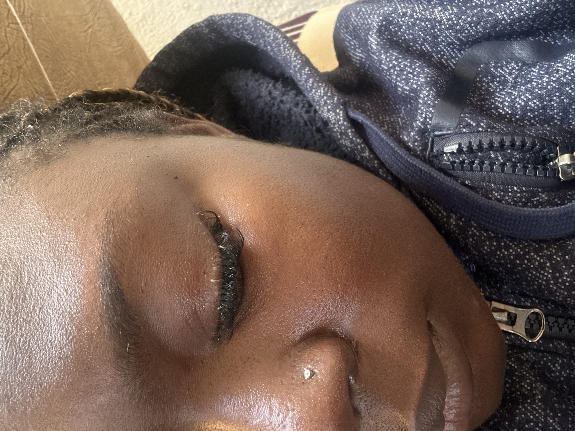 The image is a close-up, eye-level, outdoor shot of a Black person wearing a navy blue hoodie. The person's left eye is closed, and they have false eyelashes. A small silver stud is visible on their nose. The person's hair is braided and pulled back. The background is blurred, showing a light-colored surface and a hint of a patterned fabric. The lighting suggests it is daytime. The image quality is good, with clear focus on the subject's face.