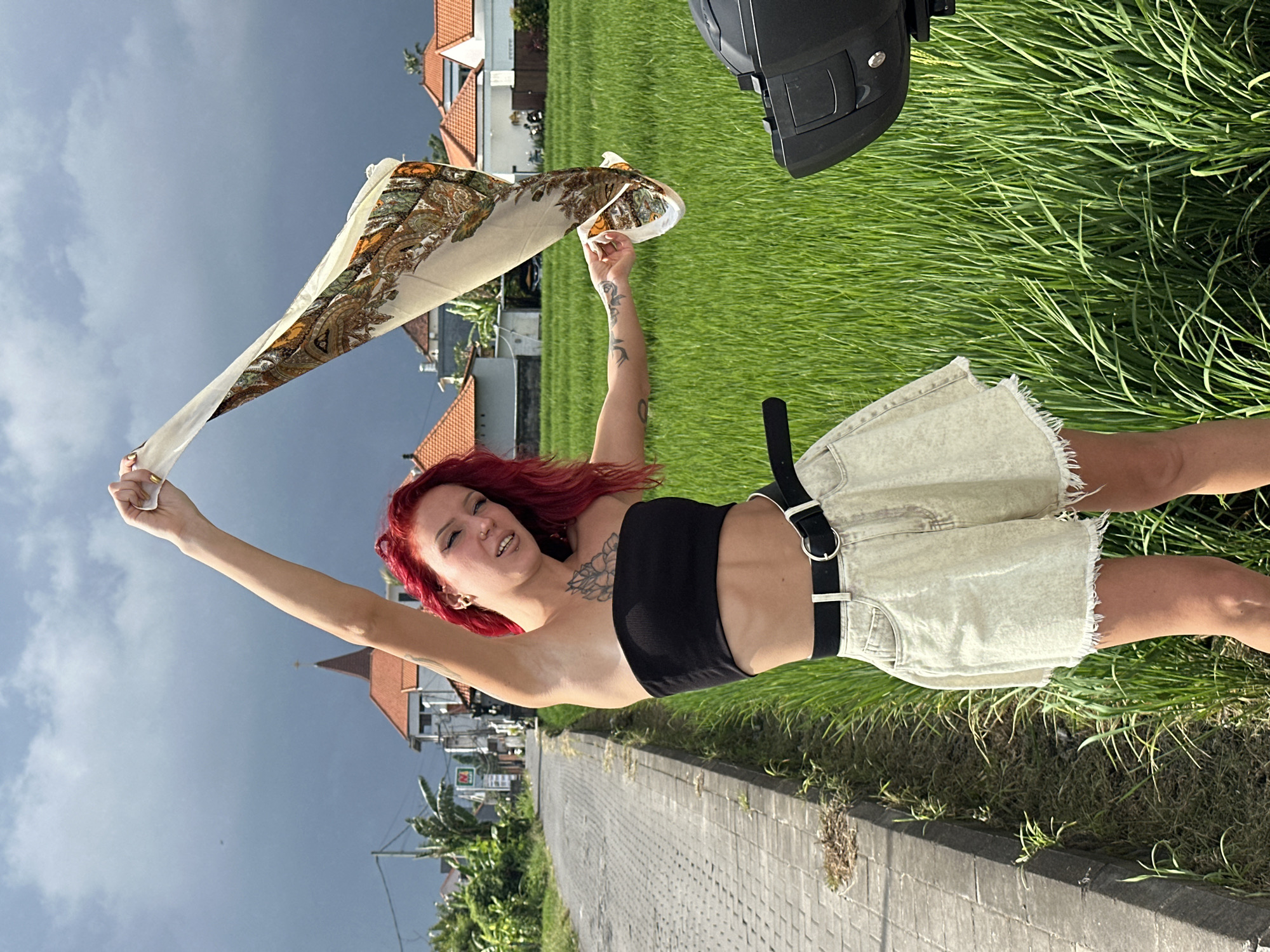 A young woman with red hair stands in North Kuta, Indonesia, on a paved roadside with a green grass field adjacent to her. She wears a black tube top and light-colored denim shorts with a black belt. Her left arm is raised, holding a light-colored patterned scarf. Tattoos are visible on her arms and chest. Buildings with red-tiled roofs and palm trees are visible in the background. The sky is overcast with dark storm clouds. A black object is partially visible at the top of the frame.