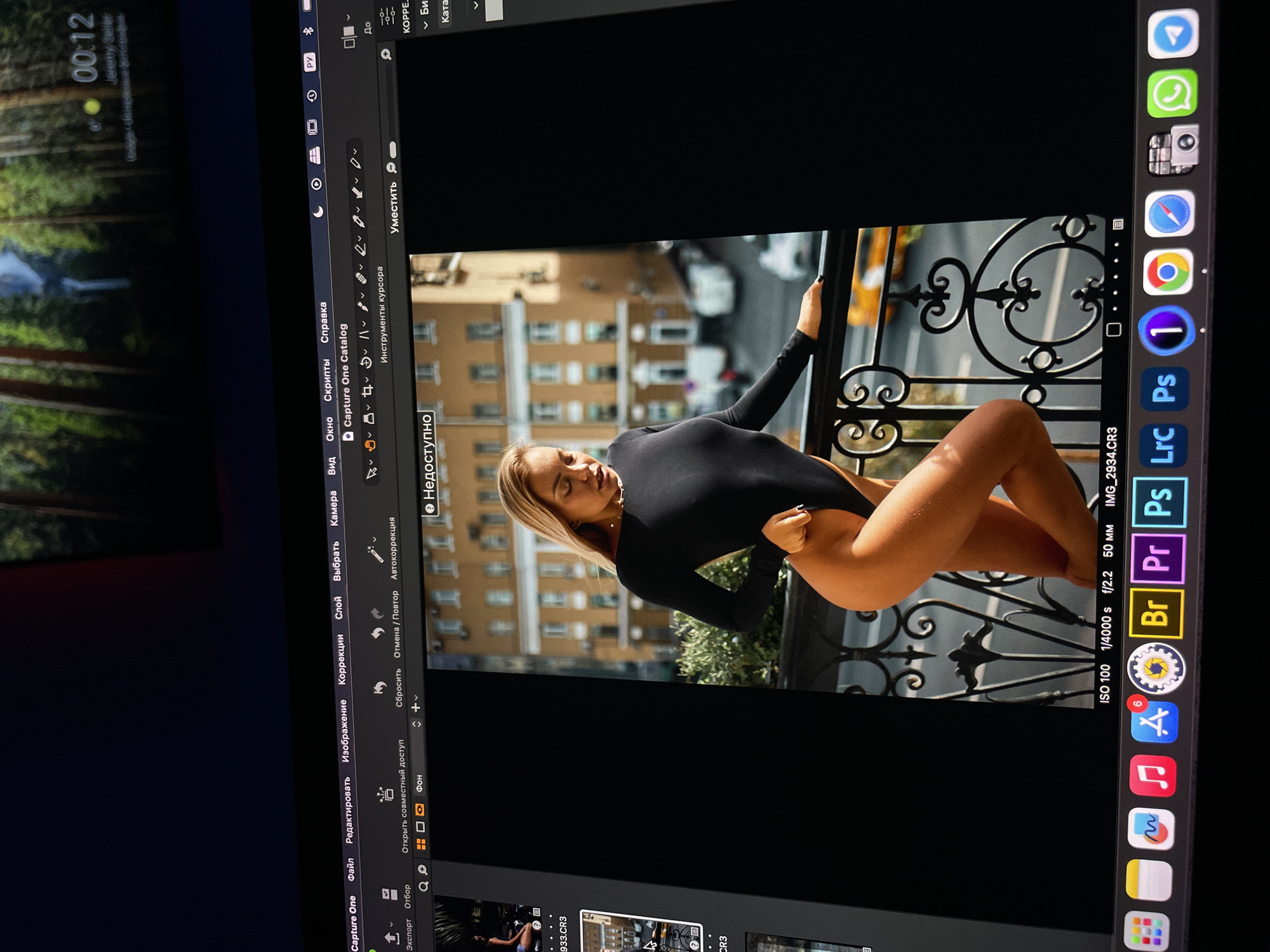 A photograph shows a woman on a balcony in what appears to be Kazan, Russia. The woman is Caucasian with blonde hair and is wearing a black bodysuit. She is leaning on the balcony railing with one hand while the other hand is out of frame. The background shows a city street with cars and buildings. The picture is displayed in Capture One editing software on what looks to be a Macbook. The ISO is set to 100, a shutter speed of 1/4000 and an aperture of 2.2mm.