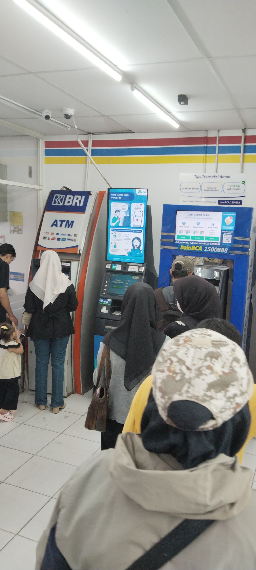 This is a vertical, eye-level, full-shot photograph taken indoors, likely in a convenience store or minimarket, given the presence of ATMs and the general lighting. The scene depicts a queue of people waiting to use ATM machines.
In the foreground, the back of a person wearing a tan hooded jacket and a camouflage-patterned baseball cap is prominently visible, facing away from the camera. To the left and slightly behind this person is another individual wearing a dark hijab and a grey long-sleeved shirt, also facing the ATMs. Further to the left, a woman in a white hijab and black jacket, blue jeans, and tan high-heeled shoes stands at the BRI ATM. She is facing the ATM, suggesting she may be either using it or about to use it. A young child in a cream-colored outfit with black trim and pink shoes stands to the far left, looking towards the woman.
The middle ground is dominated by three ATM machines. The leftmost ATM is branded "BRI" and "ATM" with various card logos. To its right is a smaller ATM with a blue screen displaying information in Indonesian, including "Tips Transaksi Aman" (Safe Transaction Tips). This screen has illustrations depicting safe ATM usage. The ATM to the right of that is a larger, blue machine, also with a screen showing Indonesian text, including "ATM SETOR TARIK" (ATM Deposit Withdraw). This screen also features icons for different transaction types. The screens of the ATMs suggest a public banking service is available here.
The background shows the interior of the establishment with bright fluorescent lighting on the ceiling. The walls are decorated with horizontal bands of red, yellow, and blue. The floor is tiled with a grid pattern. Two security cameras are visible mounted on the ceiling towards the top left of the image.
The overall impression is a mundane scene of everyday financial transactions occurring in a well-lit, commercial indoor space. The presence of multiple ATMs and the queue indicate a busy location. The specific location within Tiparraya, Indonesia, cannot be determined solely from the visual information.