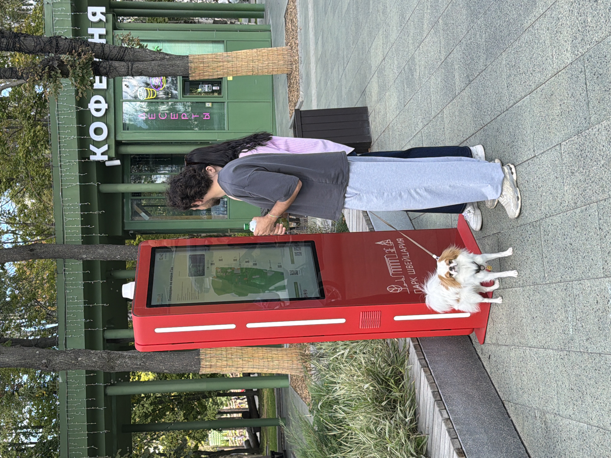 The image shows a bright red interactive kiosk displaying a map and information in Russian, likely in a park or public square. The kiosk has text at the bottom that reads "ПАРК ШВЕЙЦАРИЯ," which translates to "Switzerland Park." This indicates the location is likely Switzerland Park in Nizhny Novgorod, Russia. Two individuals are standing near the kiosk, one of them a male with curly hair wearing a grey t-shirt and light blue sweatpants, and the other a female with long dark hair wearing a pink striped shirt. They appear to be looking at the information on the kiosk. A small white and tan dog with a red leash is standing in front of the kiosk. In the background, there is a green building with "КОФЕЙНЯ" (cafe) written on it in large white letters, and some trees and greenery. The ground is paved with gray interlocking tiles. The overall scene depicts a casual outdoor urban setting on a partly cloudy day.