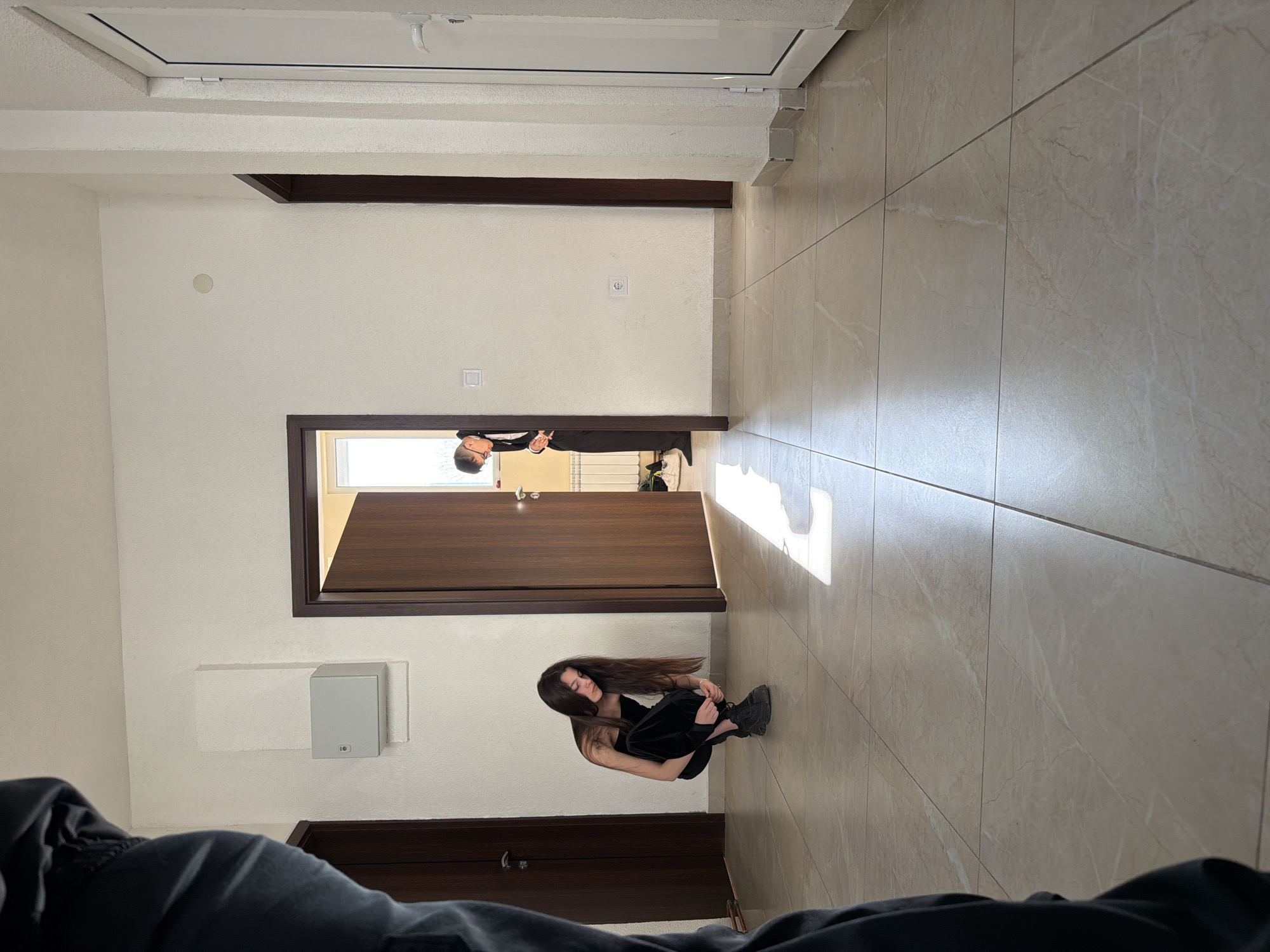 The image appears to be taken in an interior space in Shumen, Bulgaria. The perspective is unusual, possibly shot from a prone position. A person in dark clothing sits on the tiled floor, knees drawn up, in the lower center of the frame. Another person in darker formal attire is seen standing at the end of a hallway or doorway, peering into a room. The walls are painted in shades of white. There are several dark-framed doorways along the walls and a white panel on the upper portion of a wall. The floor is covered in large rectangular tiles. Light streams through a doorway casting a bright rectangular shape on the floor. The foreground is filled with a large, unidentifiable object that is black in color.