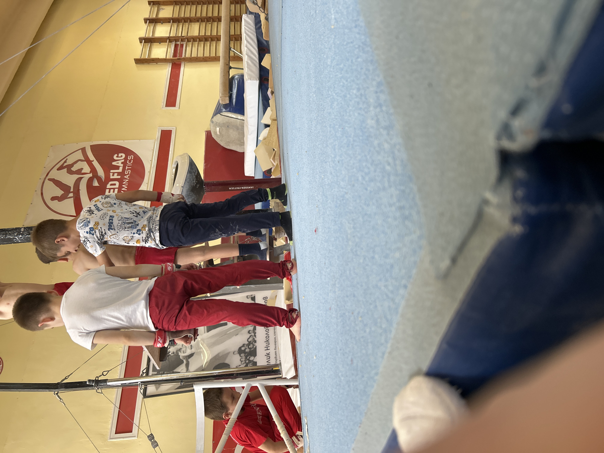 This image captures an indoor gymnastics training session in what appears to be a gym in Sofia, Bulgaria. The setting features light yellow walls, a blue padded floor mat, and various pieces of gymnastics equipment.
Several young boys are present, seemingly engaged in practice. One boy, visible from the back, wears a white t-shirt with colorful patterns and dark blue pants. Another boy in the foreground wears a white t-shirt and red pants, with wrist wraps suggesting active participation in training. Parts of other individuals are also visible, including a bare-chested boy and one in a red t-shirt in the lower right, implying multiple athletes are present.
The gym is equipped with apparatus such as parallel bars, part of which is in the immediate foreground, and what looks like a pommel horse or similar padded equipment (blue and white) further back. A red mat or block branded with "JANSSEN-FRITSEN" is also visible. Against the yellow wall, a wooden wall-mounted ladder or set of bars can be seen.
Notable visual cues include a large banner on the wall featuring a gymnast's silhouette and the text "RED FLAG GYMNASTICS". Partially obscured Cyrillic text on a poster or banner reads "елик Николо" and "първият българин, спечел", suggesting a tribute to a Bulgarian gymnast, possibly "Георги Николов, първият българин, спечелил..." (the first Bulgarian to win...). The branding "REBEL" is also visible on some equipment or clothing. The image is taken from a slightly tilted, low-angle perspective, emphasizing the floor and the base of the equipment.