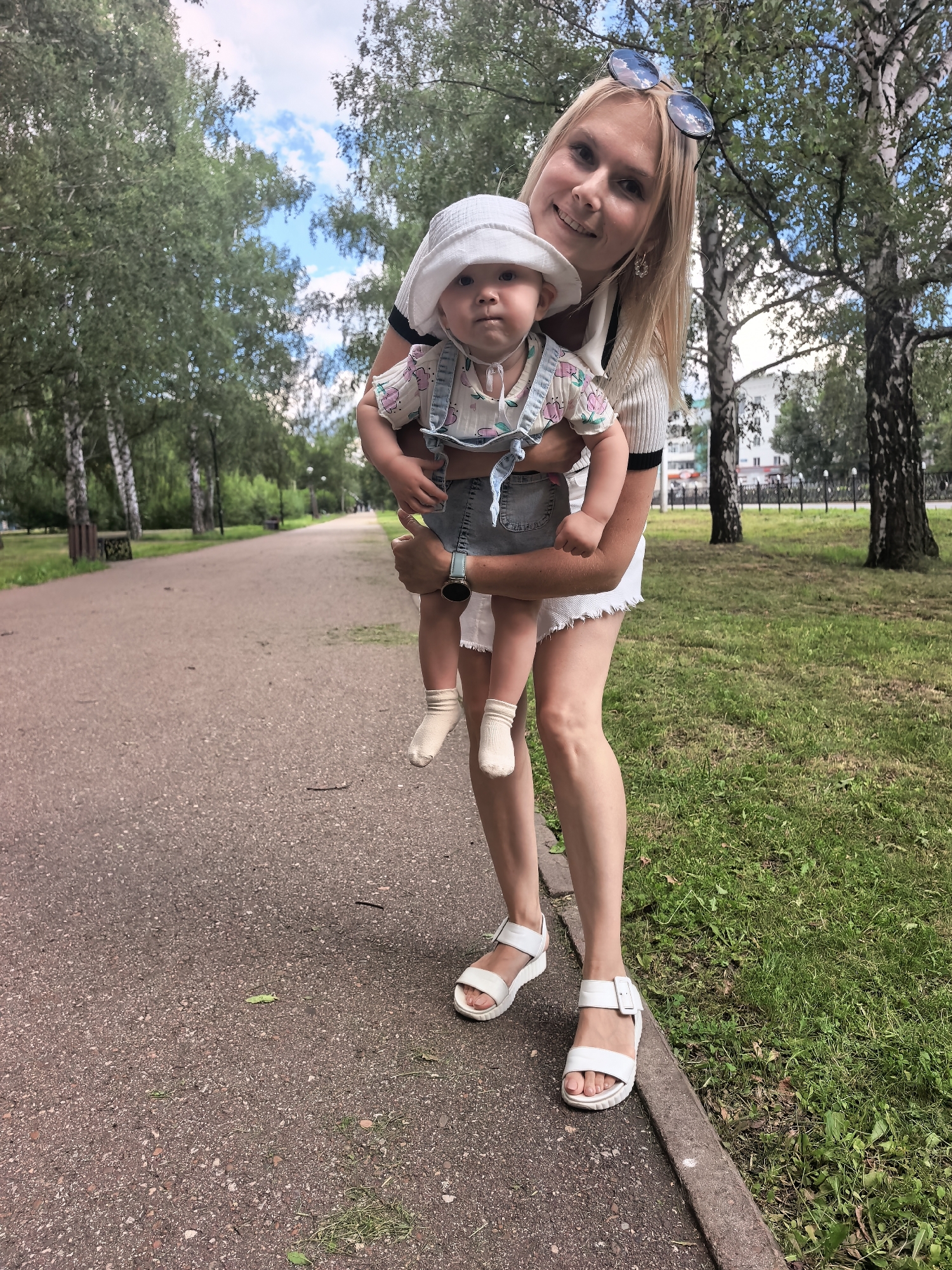 In Sterlitamak, Russia, a woman with blonde hair and sunglasses on her head is holding a baby in a park. The woman is wearing a white top and white shorts, and white sandals. The baby is wearing a white hat, a short-sleeved shirt, and denim overalls. They are standing on a paved path next to a grassy area with trees. The path appears to be a walkway in a park or public area, with benches visible in the background. The overall scene depicts a casual outing in an urban park.
