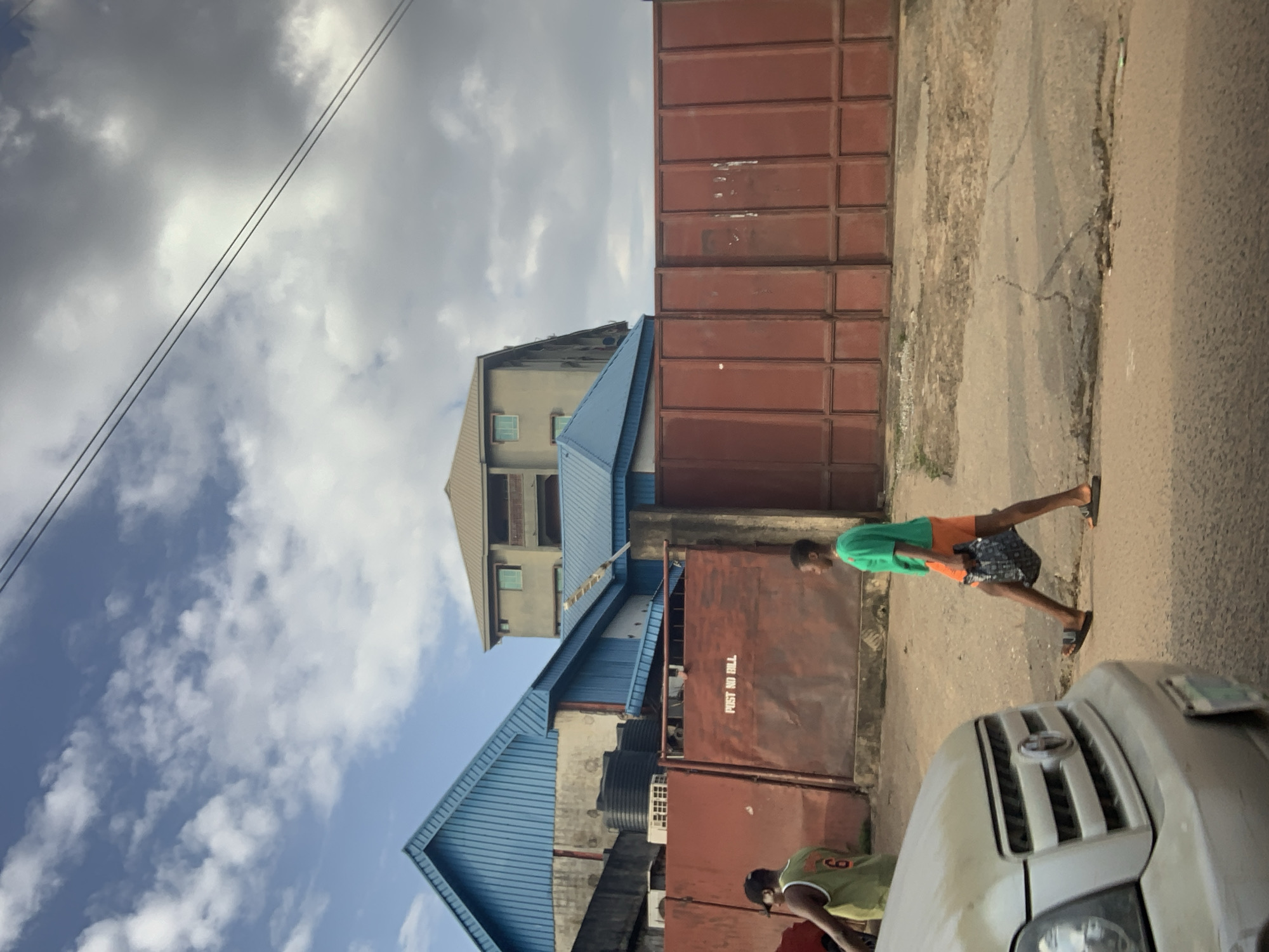 In Nkpor, Nigeria, on a street, a person wearing an orange shirt and shorts walks on the right side of the road carrying a bag. A car is parked on the left side of the road. A red metal fence spans the background. Buildings with blue and tan roofs are visible behind the fence. The sky is cloudy.