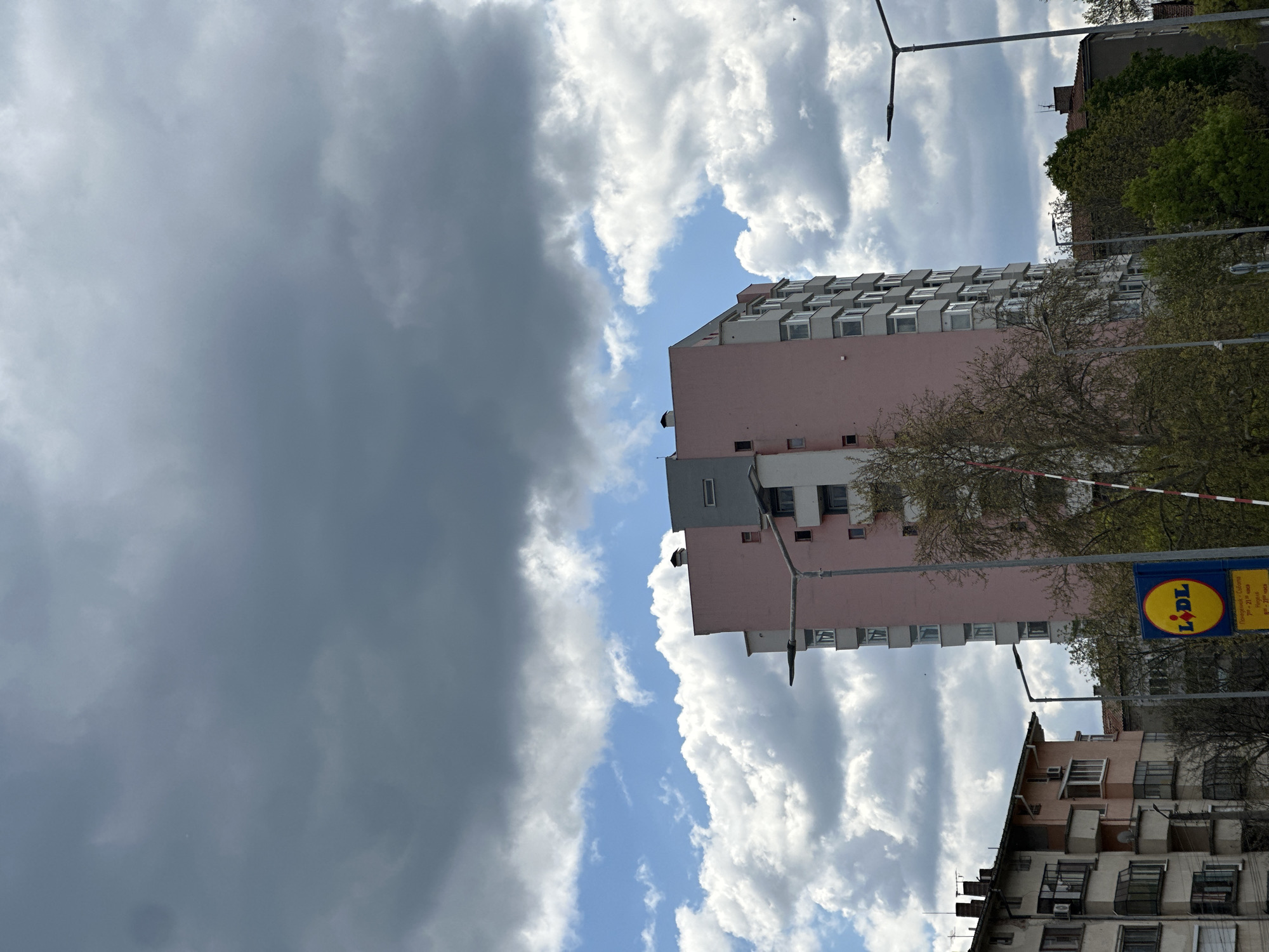The image, taken in Lovech, Bulgaria, captures an urban street scene under a dramatic sky. The upper left portion of the frame is dominated by heavy, dark grey storm clouds, while the lower right reveals patches of bright blue sky interspersed with fluffy white cumulus clouds, indicating dynamic and contrasting weather conditions.
In the mid-ground, a tall, multi-story residential building with a light pink or peach facade stands prominently. It features numerous rectangular windows, many of which appear to be enclosed balconies, and a distinct grey section near its top. To the lower right of this building, an older apartment block with varied architectural elements and visible air conditioning units can be seen. Sparse trees with light foliage are scattered throughout the scene, particularly around the buildings.
Several street light poles with their characteristic bent arms are visible, along with a partial view of a red and white striped barrier in the foreground. A yellow and blue circular sign for the "LIDL" supermarket is clearly visible, attached to a pole. Below the LIDL logo, Cyrillic text provides the store's opening hours: "Понеделник - Събота: 7:30 - 21:00 часа" (Monday - Saturday: 7:30 - 21:00 hours) and "Неделя: 8:00 - 21:00 часа" (Sunday: 8:00 - 21:00 hours). The scene depicts a static daytime environment with no people visible or specific activities taking place.