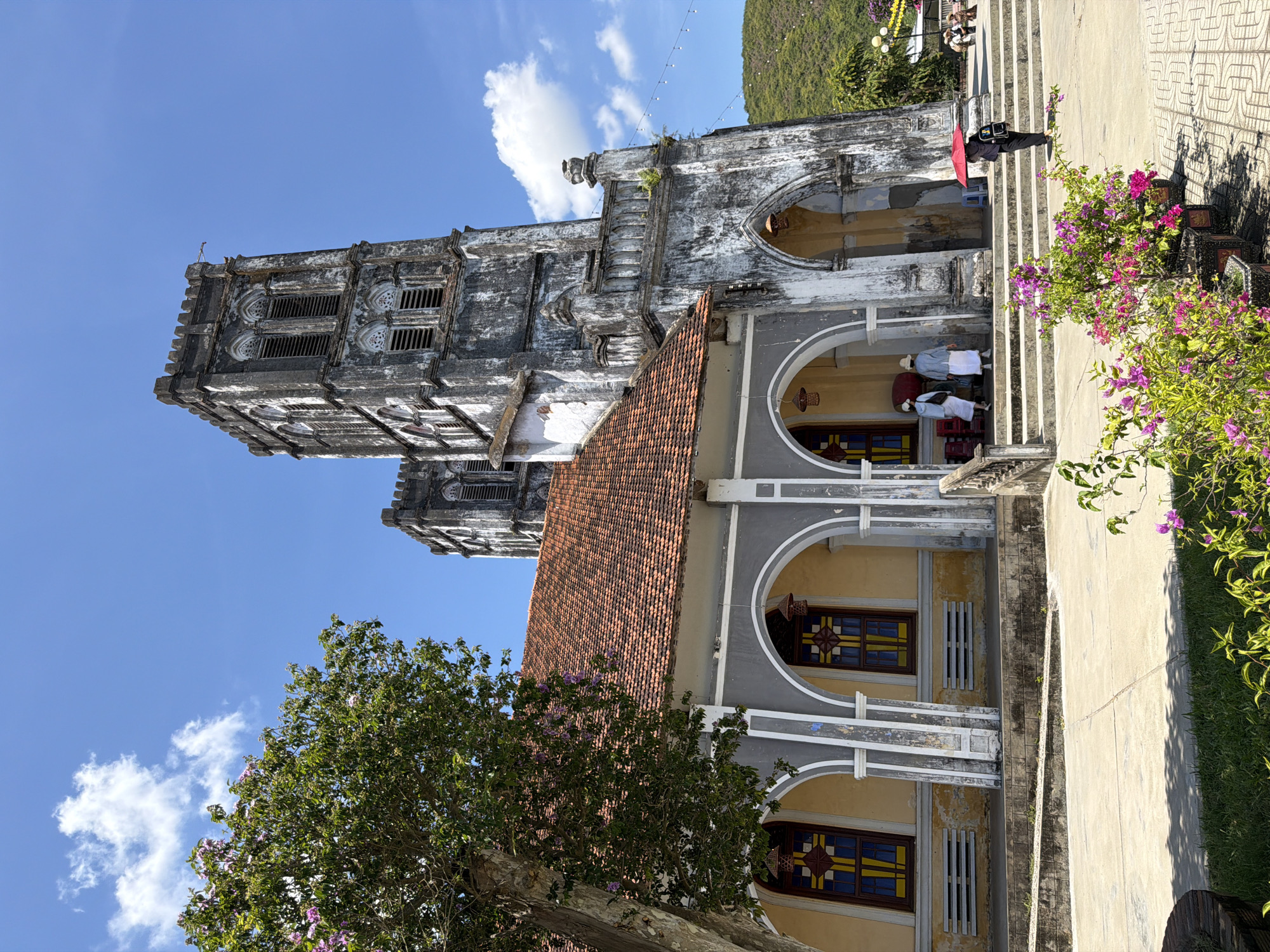 The image depicts the exterior of a stone church in Hội Tín, Vietnam, under a clear blue sky with scattered clouds. The church features a central section with a red tile roof, flanked by two weathered stone towers. Arched windows with stained glass are visible on the side of the main building.
In the foreground, a lush green tree with pink blossoms stands to the left, and pots of vibrant pink and purple flowers are arranged in front of the church entrance. Several individuals are present. Two people wearing white robes and hats stand near the entrance of the church. Another person, holding a red umbrella, walks away from the camera towards the steps of the church. In the background, more people are seated on a bench, and string lights are visible. The landscape behind the church is characterized by green, rolling hills. The overall scene suggests a bright, sunny day at a historic religious site.