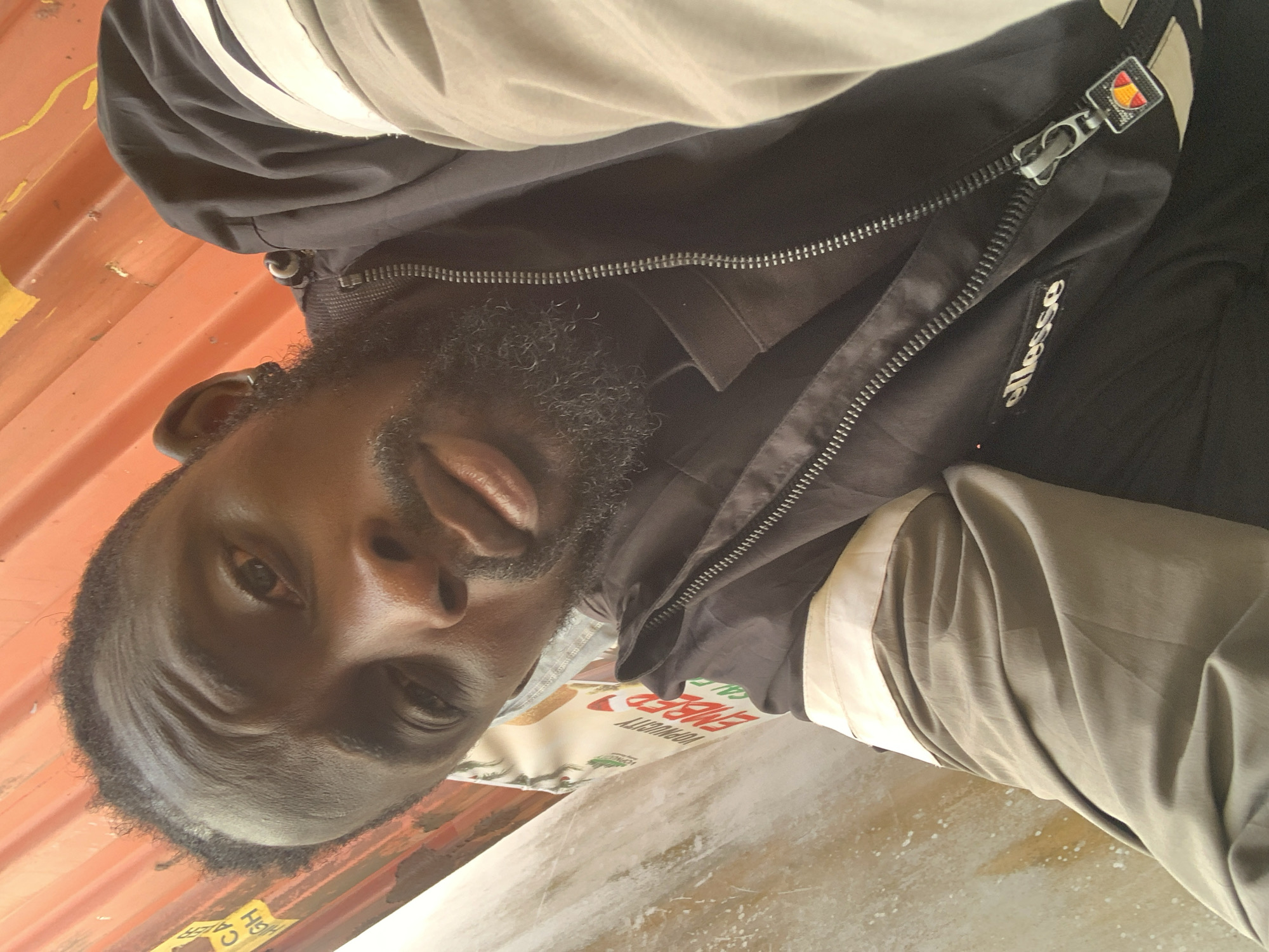 A close-up, rotated portrait shot captures a man, possibly in Lekki, Nigeria, looking directly at the camera. He has dark skin, a full, dark beard, and short dark hair. He wears a black jacket featuring lighter beige or off-white stripes and panels. A white "Ellesse" logo is visible on the left chest area, and the jacket's zipper pull also prominently displays the distinctive red and orange Ellesse logo.
The background primarily consists of a bright orange corrugated metal surface, possibly a shipping container or an industrial wall, behind his head and to his left. Below him and to his right, a lighter, textured surface, possibly concrete or a light-colored wall, is visible. An object beneath his chin, possibly packaging or a bag, shows partial text including "YOURORITY" and "ENERGY". A label with the text "HIGH C" is partially visible at the bottom left corner. The natural, clear lighting suggests the photo was taken during the daytime. The man's expression is neutral and direct, creating an engaging and casual portrait.