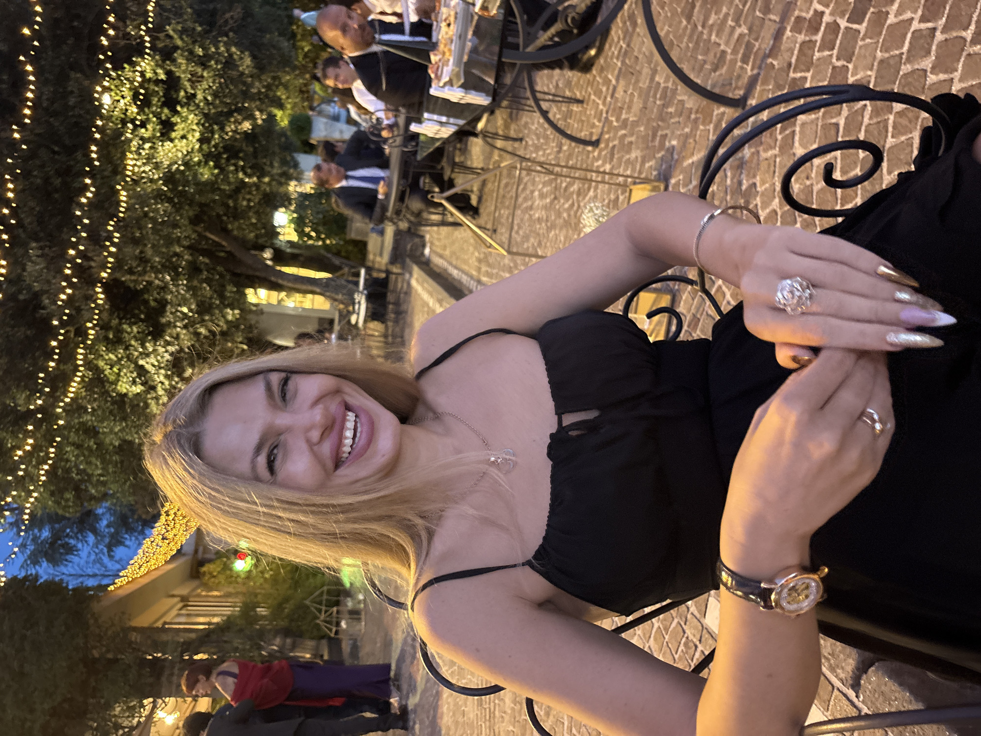 A blonde woman in a black dress is seated at a wrought iron table in an outdoor restaurant setting, likely in Castel Gandolfo, Italy, given the visible architecture and decorative lights. She is smiling and looking at the camera. Her hair is long and blonde, and she is wearing a necklace and a watch on her left wrist. She also has rings on her fingers. In the background, other diners are visible at tables, some of whom are wearing formal attire. Trees and decorative string lights illuminate the scene. The ground is paved with cobblestones.