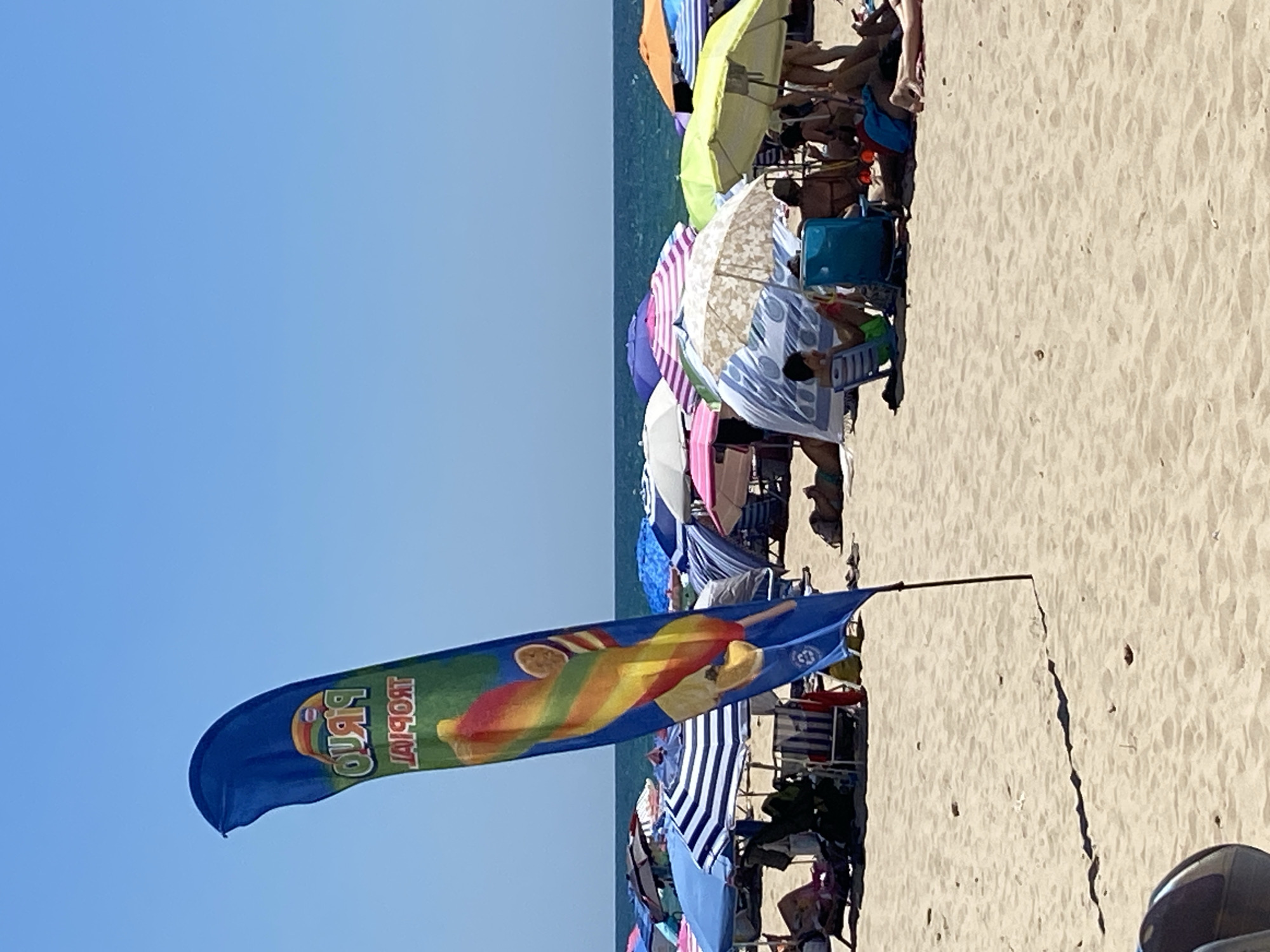 The image depicts a crowded beach in Conil de la Frontera, Spain, on a clear, sunny day. In the foreground, a blue flag with a rainbow-colored popsicle graphic and the word "TROPICAL" written on it waves from a pole. The beach is filled with numerous beach umbrellas and chairs, providing shade for beachgoers. Several people are visible, either sitting under umbrellas, lying on towels, or walking on the sand. The ocean is visible in the background, with a clear horizon. The overall scene conveys a typical summer day at a popular beach destination.