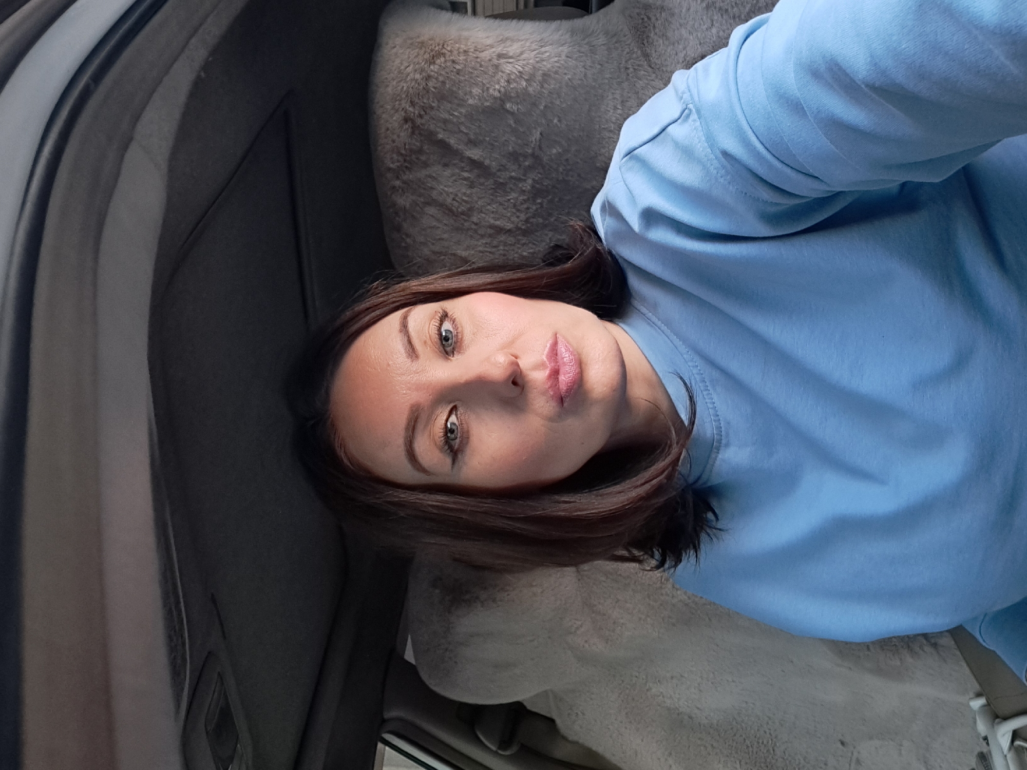 A woman with dark brown hair and light blue eyes is sitting in the driver's seat of a car. She is wearing a light blue sweatshirt. The interior of the car is visible, including the headliner, sunroof, and fuzzy grey seat covers. The woman is looking directly at the camera with her lips slightly pursed. The scene appears to be a selfie taken inside a vehicle. There is no information to suggest the location is the city of Irkutsk, Russia.