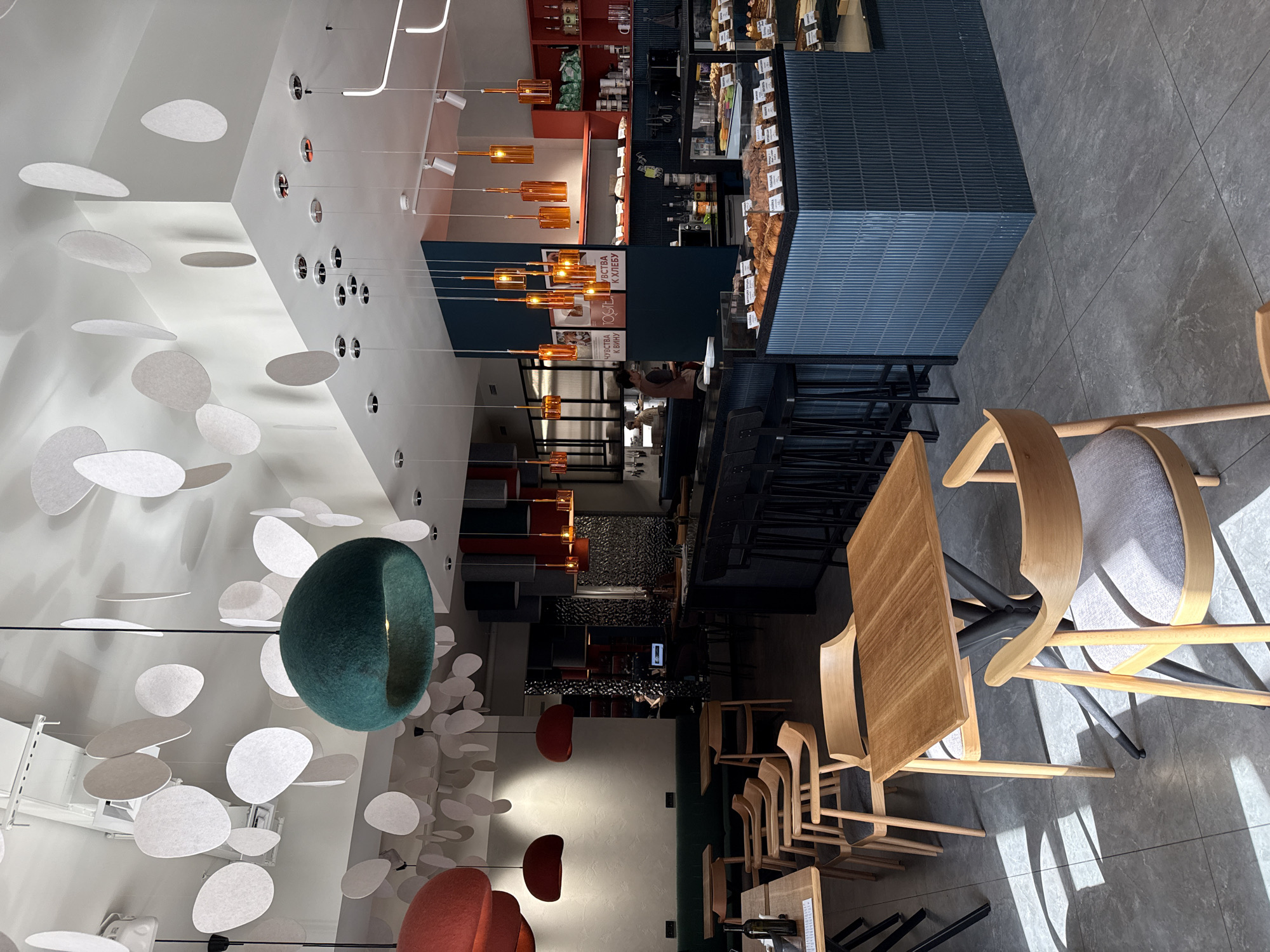The image shows the interior of a modern cafe or restaurant. A prominent design feature is a ceiling installation consisting of numerous white, disc-shaped elements and a large, green, donut-shaped object. The restaurant has a counter with a display of food items, likely baked goods or pastries. Tables and chairs are arranged for customers. The lighting is a combination of recessed ceiling lights and hanging fixtures with warm-toned bulbs. There is a mix of seating options including wooden chairs with grey cushions. In the background, there are shelves with various items, possibly ingredients or merchandise. The floor is tiled in a neutral color. Based on other images from a search engine the image is from the FORNO cafe located in Krasnoyarsk, Russia.