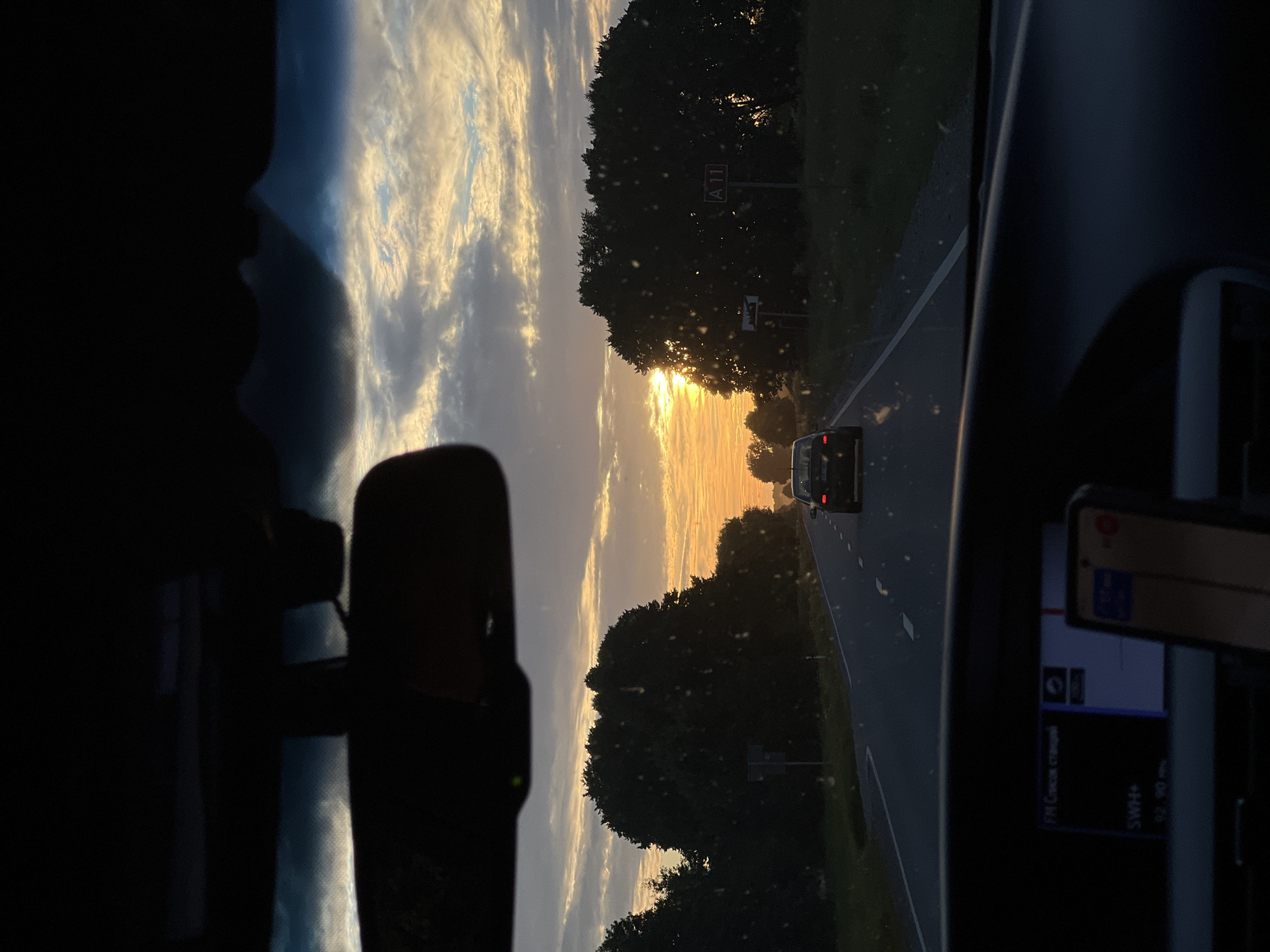 The image shows a view from inside a car driving on a road towards a sunset. The road is bordered by trees on both sides. There is another car in front of the viewer's car on the same road. The sky is filled with clouds and the setting sun is visible, casting a warm glow. In the foreground, the dashboard of the car is visible, with a car stereo and a phone mount with a phone. There are road signs visible on the side of the road, one of which appears to be a route marker for A11. The scene is likely taking place in or near Nīca, Latvia, given the mention of the A11 highway which runs through the area. The overall atmosphere is calm and serene, typical of a sunset drive.