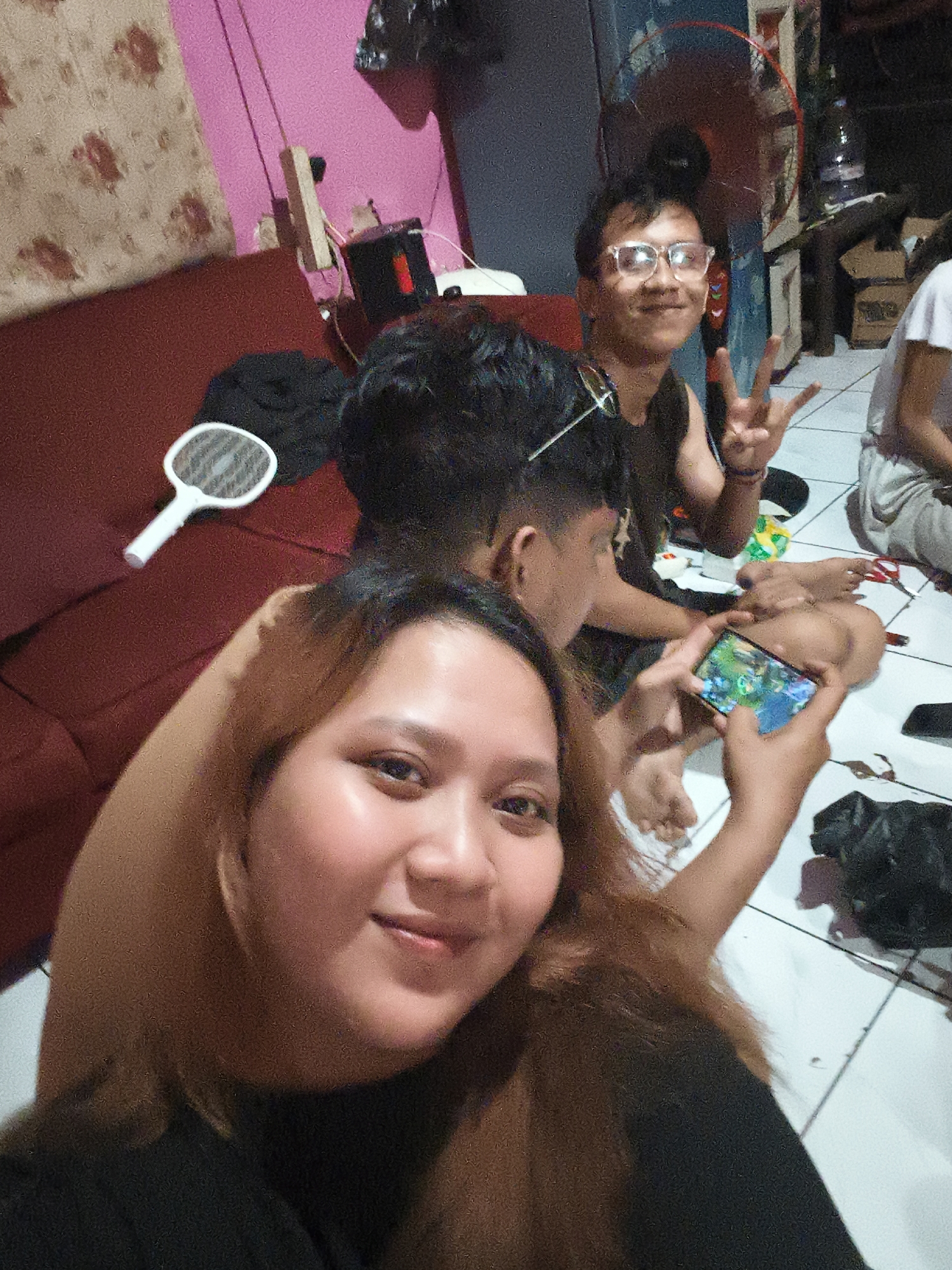The image depicts an indoor scene, likely a living room or common area, with several individuals gathered. The flooring appears to be tiled. In the foreground, a young woman is taking a selfie, holding a smartphone in her right hand, which displays a game. Her left arm is bent and resting on her head. To her left, a white electric mosquito swatter lies on a maroon sofa. Behind her, other individuals are visible: a young man wearing glasses is in the middle ground, making a peace sign with his right hand and smiling at the camera. To his right, another person's back is visible, and further back, another individual is seated on the floor. The room's walls are painted pink and have a patterned wallpaper. A standing fan is visible in the background. A black trash bag is on the floor to the right. The lighting suggests it is evening or nighttime. The presence of the mosquito swatter indicates this is likely a residential area in Depok, Indonesia, where such pests are common.
