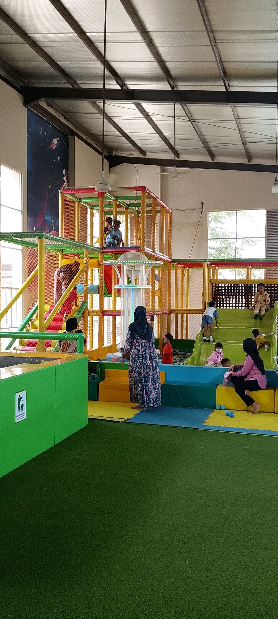 The image depicts an indoor children's play area, likely at "KidZania" in Sriamur, Indonesia, as suggested by the context. The space is characterized by a large, multi-level soft play structure with vibrant yellow, red, and green netting and padding. Children are visible engaging in various activities within this structure, including playing and climbing. To the left, a large green barrier with a "no littering" sign is present, bordering a green carpeted area. In the foreground and mid-ground, several adults and children are interacting. An adult in a floral dress is standing near the play structure, observing. Another adult in a pink hijab is seated on a yellow and blue padded platform, with a child in pink beside them. A young boy in a red shirt stands to the left of the adult in the floral dress. The overall environment is brightly lit, with a high ceiling featuring industrial beams and translucent panels, suggesting it is part of a larger building or warehouse. The flooring is a combination of green carpet and multi-colored padded mats.