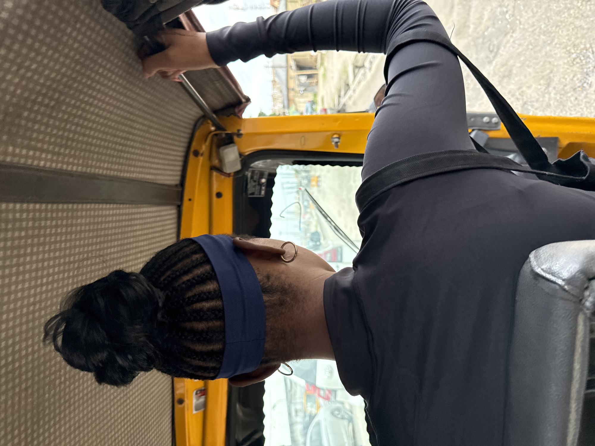 The image captures an outdoor scene from the interior of a yellow auto-rickshaw in Ado, Nigeria. The perspective is from behind a person seated in the passenger area, facing forward. The person has dark skin and their hair is styled in braids pulled up into a bun, adorned with a dark blue headband. They are wearing a black long-sleeved shirt. Their left arm is raised, with their hand resting on the auto-rickshaw's roof, while their right arm is down, visible across their back. A black strap, possibly from a bag, is seen crossing their back. The interior of the auto-rickshaw features a textured, light-colored ceiling. Through the windshield, the street is visible, with traffic including other vehicles and a glimpse of buildings and vegetation in the background. The overall impression is of a person traveling within the city.