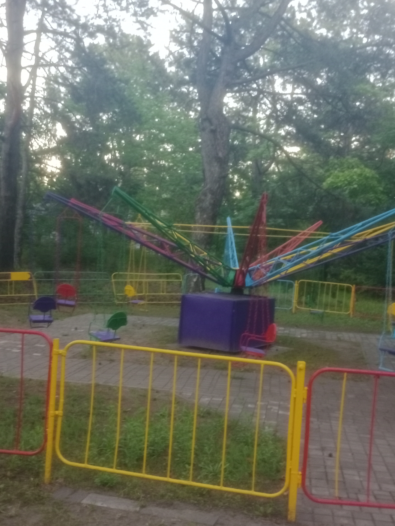 The image depicts an outdoor amusement park area, likely a children's playground, in Horishni Plavni, Ukraine. In the center of the frame is a multi-colored amusement ride featuring a central purple base and several colorful arms extending outwards, each equipped with a seat. Some of the seats are visible, including red, green, and purple ones, some appearing to be in motion or ready for use. The ride is surrounded by yellow and red metal safety fences. In the background, tall trees with dense green foliage suggest a park or wooded area. The ground appears to be paved with bricks or tiles in some areas and covered with grass in others. The lighting suggests it is daytime, possibly overcast or late afternoon, as the sunlight is diffused through the trees. No people are visible in the image.