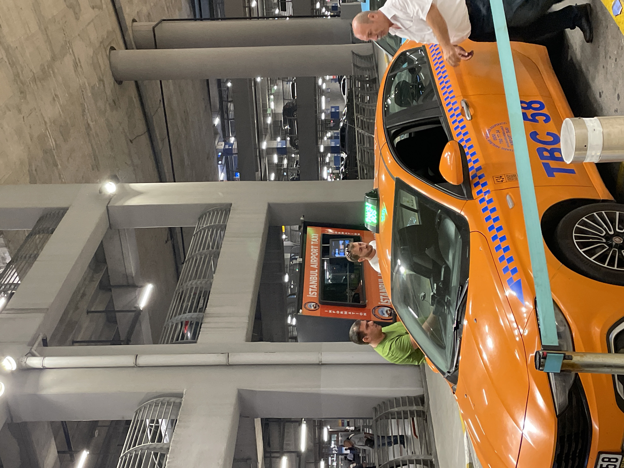 An orange Istanbul Airport Taxi is parked in a concrete parking garage in Arnavutköy, Turkey. The taxi has blue checkered patterns on its doors and the number "TBC 58" written in large blue letters on its side. Two men are standing near the taxi, one wearing a green shirt and the other a white shirt. A third man, wearing a white shirt and dark pants, is walking away from the taxi. An orange information booth with "ISTANBUL AIRPORT TAXI" written on it is visible behind the taxi. The garage has exposed concrete pillars and a concrete ceiling.