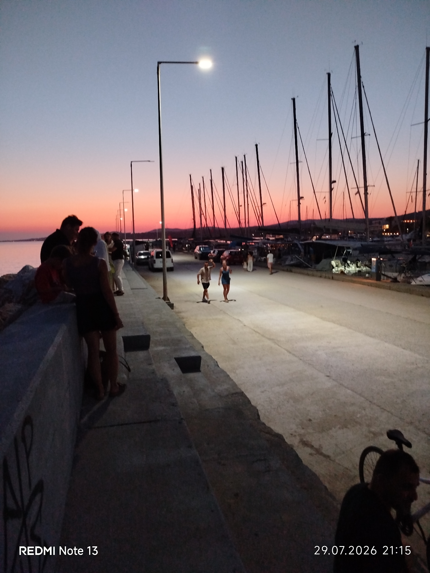 The image captures a twilight scene at a harbor in Sithonia Municipal Unit, Greece. The sky displays a gradient of pale blue and soft orange hues, indicative of sunset or sunrise. A paved walkway runs parallel to a harbor filled with numerous sailboats, their masts creating a striking silhouette against the sky. Several streetlights are visible along the walkway, illuminating the path.
On the left side of the image, a group of people are gathered near a stone wall overlooking the water. One individual is partially visible, while others are more clearly defined, suggesting social interaction. Further down the walkway, two individuals are engaged in activities, one appearing to be skateboarding or rollerblading. In the background, several vehicles, including cars and vans, are parked.
Towards the right side of the walkway, another individual is seen near a bicycle. The overall atmosphere suggests a public gathering space, possibly a promenade or waterfront area, frequented by both locals and visitors enjoying the evening. The presence of sailboats and the architectural style of the buildings in the distance point towards a coastal town or resort.