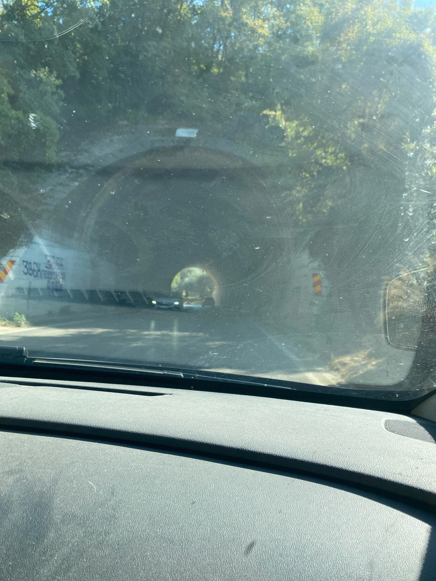 The image shows a view through a car windshield looking into a tunnel entrance. The tunnel is dimly lit, with a small circular opening visible at the far end, revealing daylight and trees. Several cars are visible inside the tunnel, heading towards the exit. On the left side of the tunnel entrance, a white sign with red and yellow markings is visible, along with some text. On the right side, a smaller sign with red and yellow markings is present. The windshield is dirty, with smudges and dust obscuring some of the view. The foreground of the image shows the textured dashboard of the car. The context provided indicates the location is in Kruče - Kërruçi, Montenegro, suggesting this is a road or highway tunnel in that area.