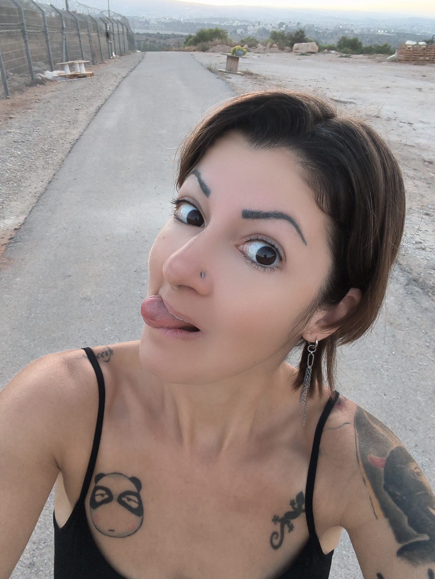 An outdoor, eye-level, medium shot shows a woman with dark brown hair, a nose piercing, and visible tattoos on her chest and arm. She is wearing a black tank top and a chain earring in her left ear. Her eyes are wide, and her tongue is sticking out. The background is an asphalt road with a chain-link fence on the left and a hilly, arid landscape with scattered rocks and vegetation in the distance. The scene is set in the Lachish Regional Council, Israel, a region characterized by its agricultural and semi-arid terrain. The time of day appears to be late afternoon or early evening, with soft, diffused light.