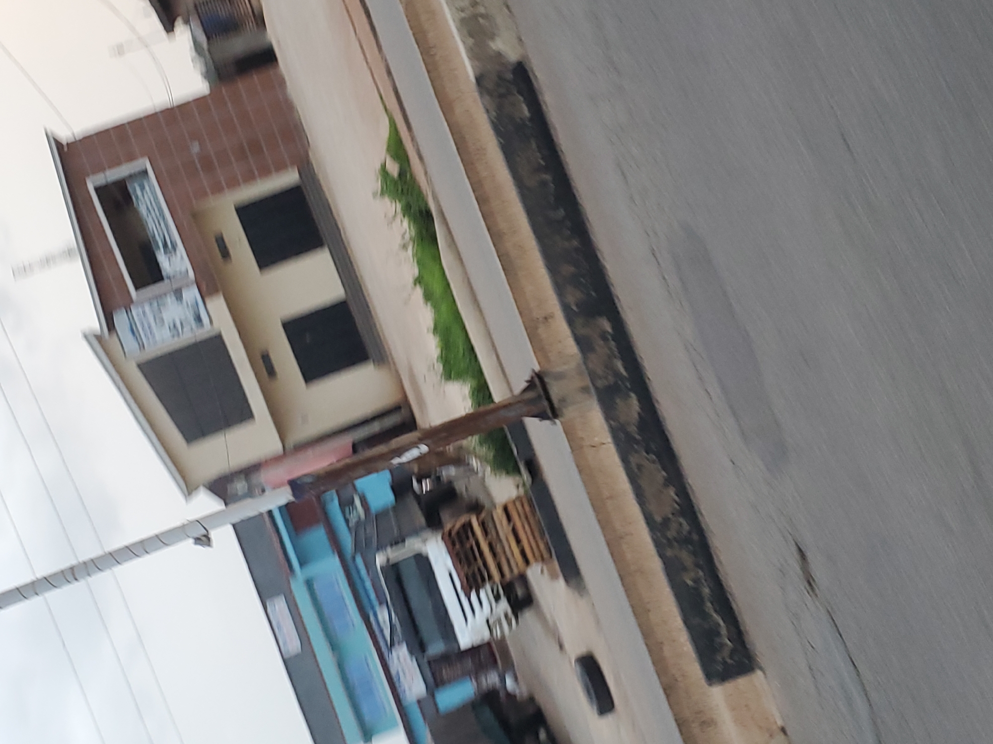 The image depicts a street scene in Gura Top, Nigeria. In the background, a two-story building with a light-colored facade and reddish-brown brick accents is visible. The building has several dark, shuttered windows and a prominent sign. To the left of this building, another structure with a blue facade and metal roofing can be seen, with a white truck parked in front of it. A stack of wooden pallets is also present near the truck. A black tire lies on the ground near the curb. The foreground shows a gray asphalt road with a concrete curb and a narrow strip of green vegetation. A light pole with overhead wires stands on the right side of the image. The sky is overcast. The image appears to be taken from a moving vehicle, indicated by the motion blur.