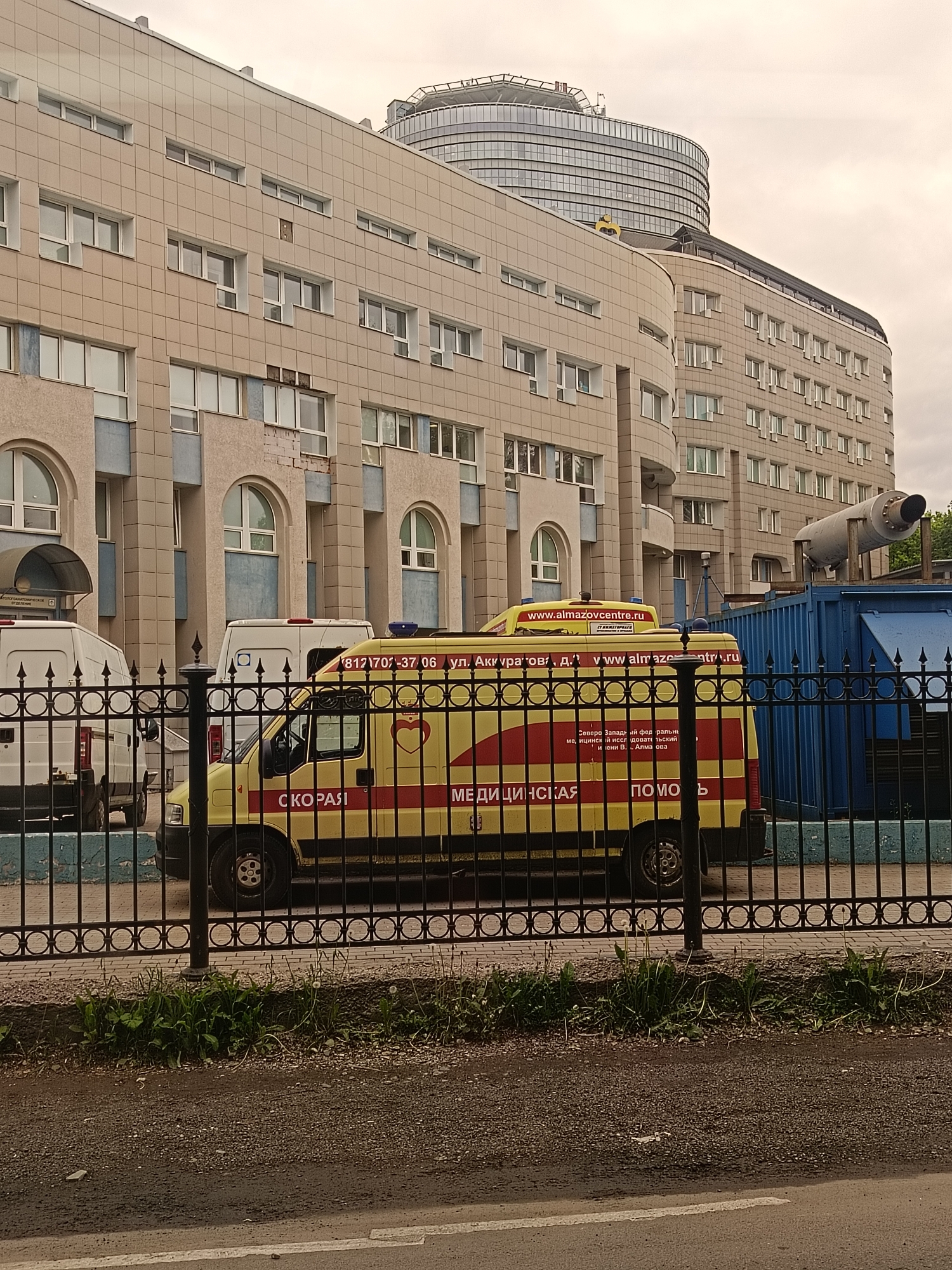 The image captures a scene in Saint Petersburg, Russia, featuring several ambulances parked outside a building. The primary ambulance is yellow with red stripes and Cyrillic writing, including the words "СКОРАЯ МЕДИЦИНСКАЯ ПОМОЩЬ" (Ambulance Medical Assistance). The side of the ambulance shows a phone number and website, "www.almazovcentre.ru", along with the street address "ул. Аккуратова, д." (Akkuratov street, building). The building is a large, multi-story structure with a beige tiled facade and arched windows on the lower levels. A modern, cylindrical building with a helipad can be seen in the background. A dark metal fence runs along the front of the scene, and a blue container and large cylindrical exhaust pipe-like structure are visible to the right of the ambulance. The foreground consists of a street with a white line and a small patch of vegetation. The sky is overcast.