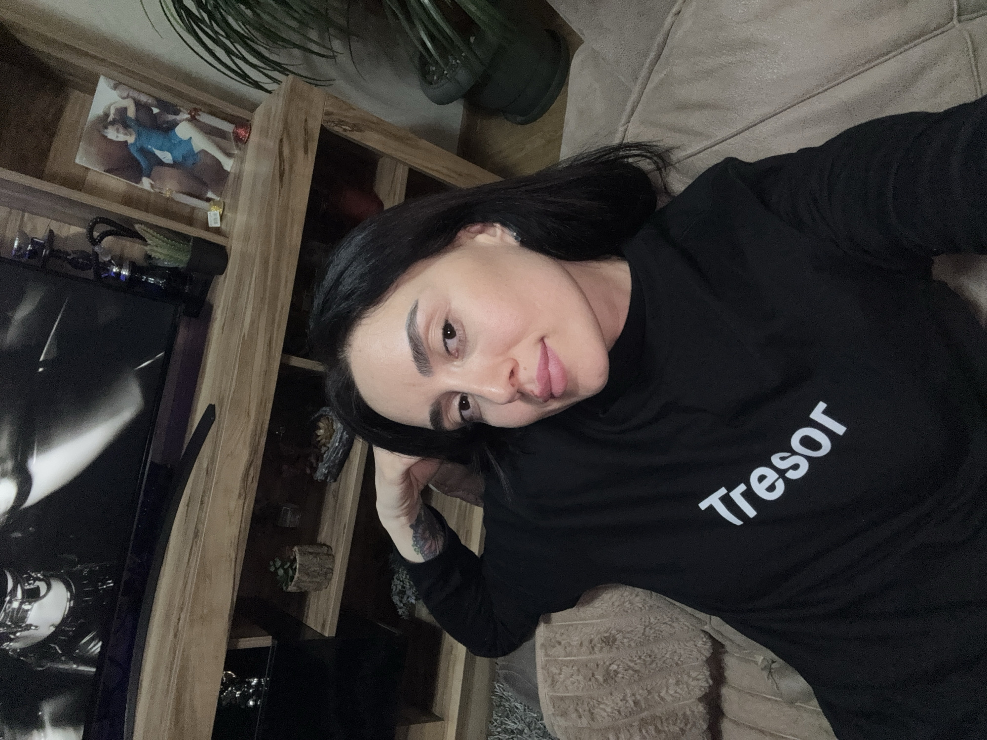 A woman is lying on a light beige sofa. She is wearing a black long-sleeved shirt with the word "Tressor" printed in white on the front. She has dark hair and fair skin, with her head resting on her hand which reveals a tattoo on her inner arm. A wooden shelving unit is visible in the background, holding various items including a photo of a woman in a blue dress, plants, and other decorative objects. A television with a black and white image displayed is to the left of the shelving unit. A large potted plant is located above the shelving unit. The environment suggests an indoor living space. There is no indication that the image was taken in Tbilisi, Georgia.