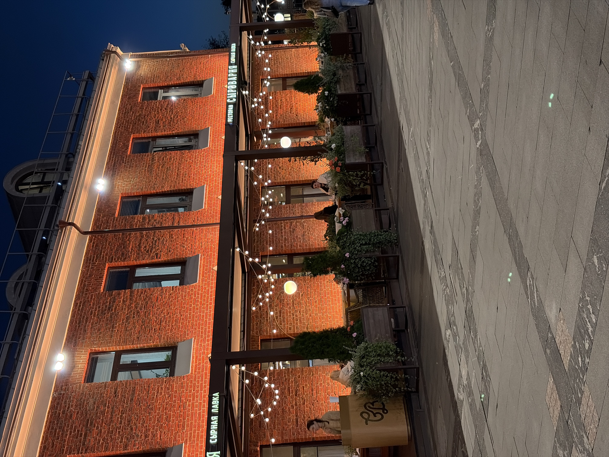 The image depicts an exterior view of a brick building at night in Moscow, Russia, featuring an outdoor dining area illuminated by string lights and exterior lighting. The building's facade is a warm orange due to upward-facing lights, and it has multiple windows. The outdoor seating area has tables, planters with greenery, and a canopy. Several individuals are visible, some seated at tables and others standing. The foreground shows a paved walkway with embedded green lights. The scene conveys a lively, atmospheric evening setting.