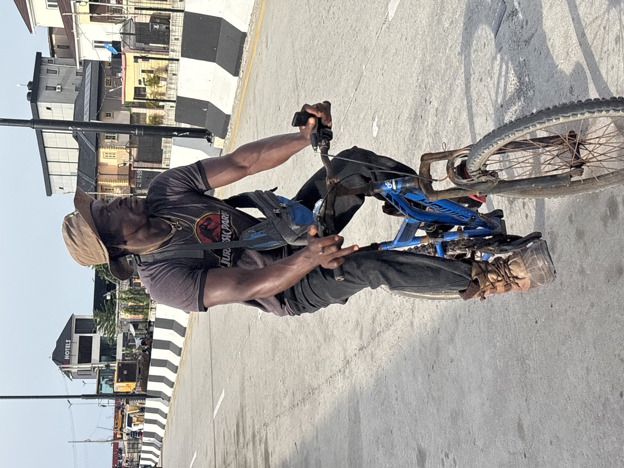 A man rides a blue bicycle along a concrete road in Igboefon, Nigeria. The man wears a khaki hat, a dark Jurassic Park t-shirt, dark pants, and brown boots. He also has a bag slung across his shoulder. His bicycle has rusty wheels. The road is marked with black and white striped barriers. Buildings are visible in the background, including one labeled "HOTELS". Road construction equipment is also present.