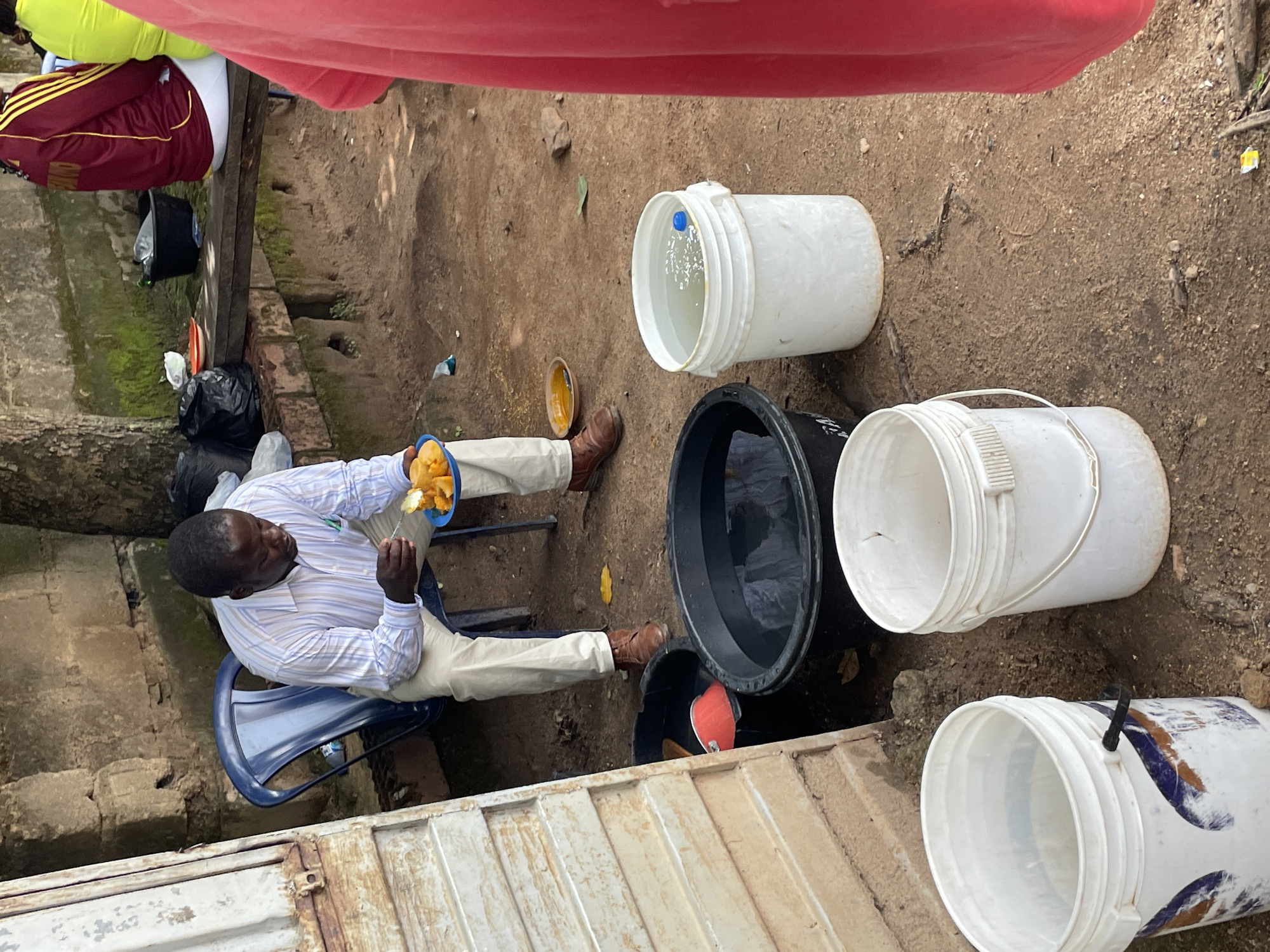 A middle-aged African man, wearing a striped long-sleeved shirt and light-colored trousers, sits on a blue plastic chair and eats from a blue plastic plate filled with sliced yellow fruit, likely mangoes. He is holding a fork in his right hand. Beside him are two large black buckets, one containing dark water and the other empty. In front of him is a white plastic bucket filled with clear water, with a blue object floating in it. In the background, there is a wooden bench on which several black plastic bags and a red and white plate are placed. Two other individuals, one wearing a red and yellow jersey and another in a bright green top, are partially visible in the background, suggesting a social gathering. The scene appears to be outdoors, with a textured dirt ground and a stone wall in the background. The overall impression is a casual, everyday moment in a somewhat rustic setting, possibly in the city of Jos, Nigeria.