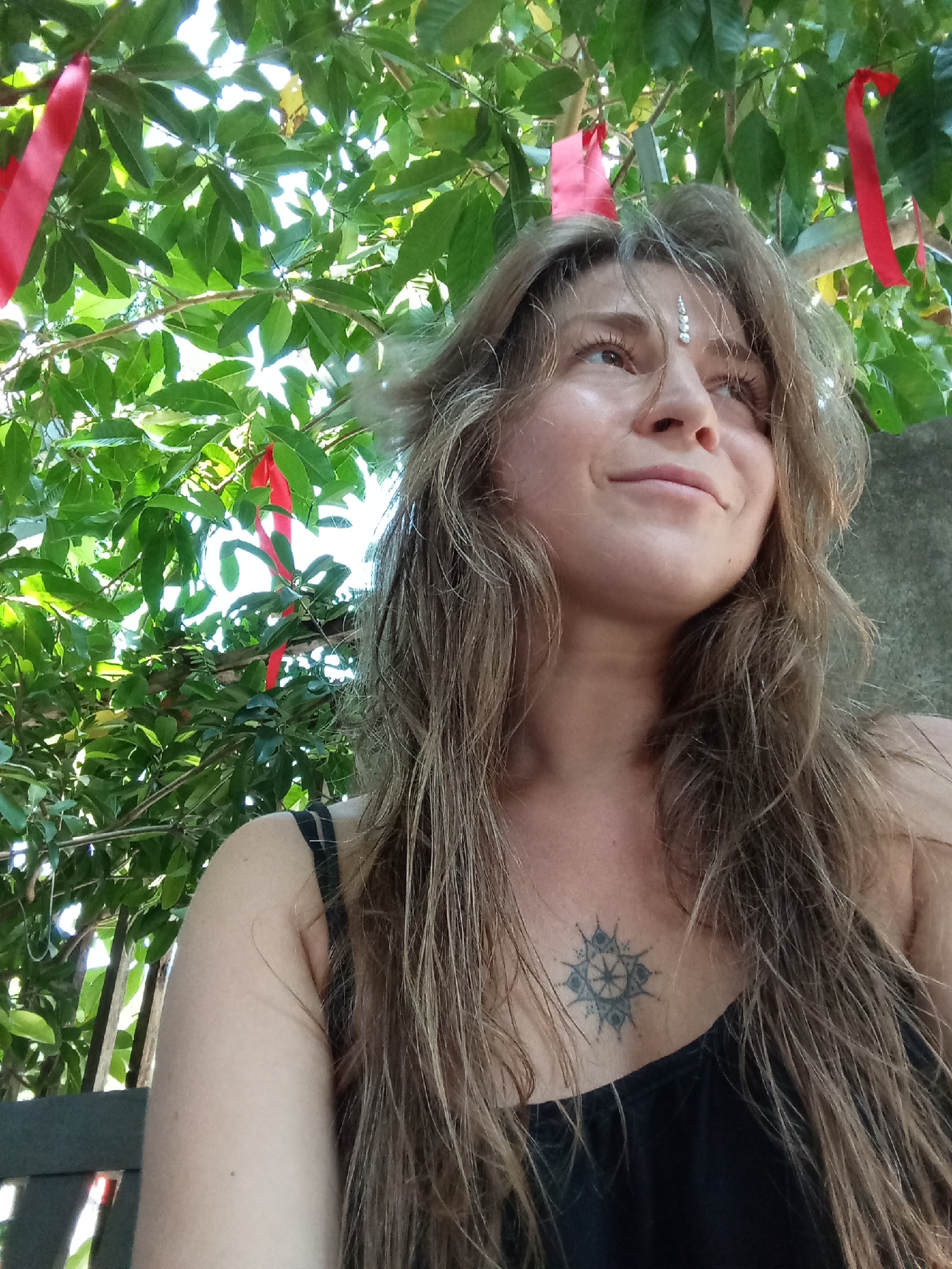 A medium shot depicts a woman with long, light brown hair, looking slightly upward and to the right. She wears a black tank top and has a tattoo on her chest. Above her are branches with green leaves, adorned with red ribbons. She has some decorative silver bindi on her forehead. She appears to be in an outdoor setting. The location could not be determined to be near Bac Natural Heritage Area Trang, Vietnam based solely on the image.