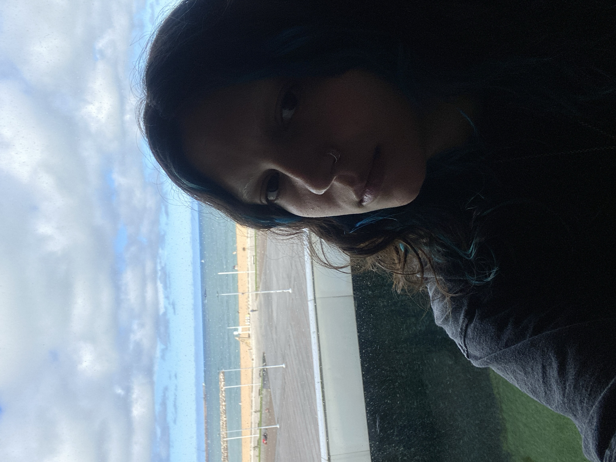 A dark-haired person with a nose ring is looking at the camera. In the background, visible through a window, there is a view of a beach, ocean, and cloudy sky. The beach has a series of light-colored posts and is bordered by a breakwater. The location is likely in Gijón/Xixón, Spain, given the prompt's mention. The person is wearing a dark-colored shirt.