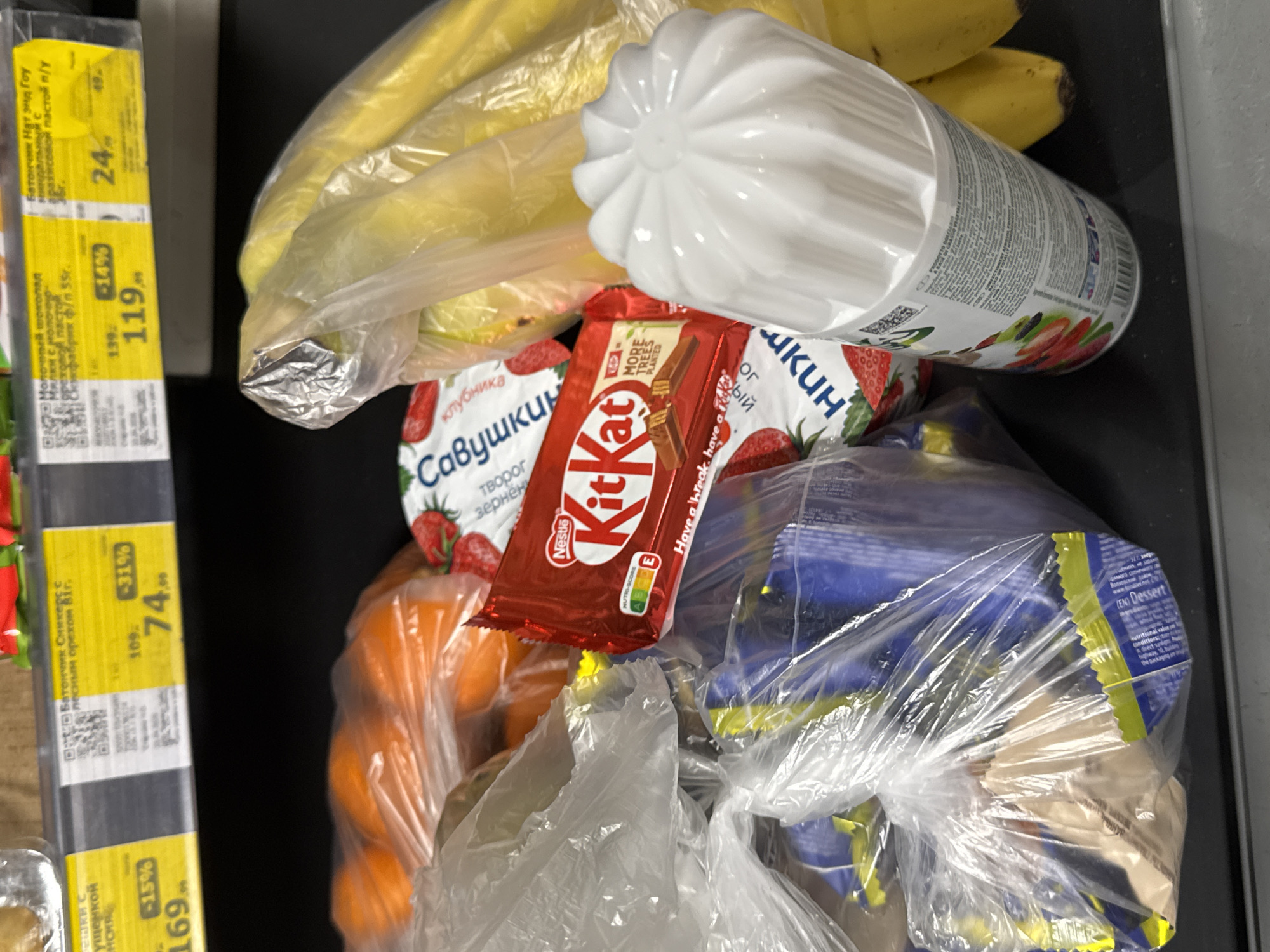 A close-up, top-down shot captures an assortment of groceries resting on what appears to be a supermarket checkout belt or counter. The items are bundled in clear plastic bags or individually packaged, suggesting a recent purchase.
Prominently featured are yellow bananas in a clear bag, a white ribbed plastic bottle (possibly a dairy drink) with Russian text and fruit graphics, and a red KitKat chocolate bar wrapper. The KitKat wrapper displays "Nestlé," "NUTRI-SCORE E," "Have a break, have a KitKat," and "MORE TREES PLANTED." Below the KitKat are two packages of "Савушкин" (Savushkin) brand dairy product, with "клубника" (strawberry) and "творог зернёный" (grained curd cheese) visible. Another clear plastic bag holds several bright orange items, likely mandarins or oranges. Towards the bottom right, a tied clear plastic bag contains multiple blue and yellow packaged items, with "EN Dessert" visible on one package.
To the left, several yellow price tags common in Russian supermarkets are partially visible. Text from these tags includes:
* "Батончик Нат энд Гоу миндальный с арахисовой пастой" (Nat & Go bar almond with peanut butter) priced at 24₽ (originally 49₽).
* "Молочный шоколад с ореховой пастой" (Milk chocolate with nut paste) priced at 119₽ (originally 139₽).
* "Батончик Сникерс с лесным орехом 81г." (Snickers bar with hazelnuts 81g) priced at 74₽ (originally 109₽).
* Another tag mentions "с сушенкой" (with dried fruit) and a price of "169₽".
The overall scene depicts a typical grocery haul at a supermarket, with the Russian text and currency indicating a location in Russia, consistent with the provided context of Perm.