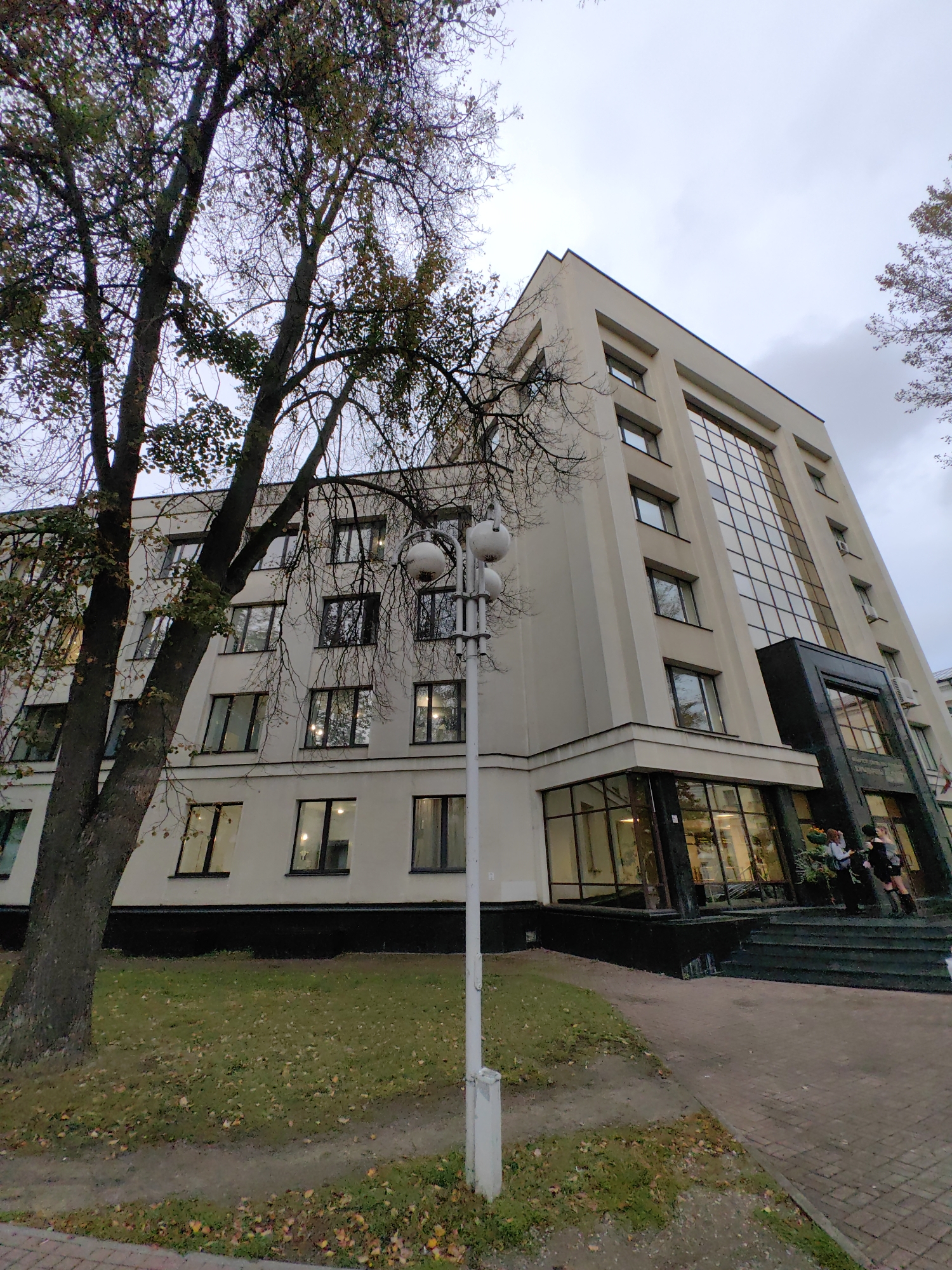 The image shows a modern, multi-story building with a light-colored facade and a large glass-paneled section on one side, located in Minsk, Belarus. A large tree with bare branches is prominent in the foreground on the left. The ground is covered with grass and fallen autumn leaves. A paved walkway leads to the building's entrance, which features steps and a dark-colored base. Two individuals, appearing to be female, are visible on the steps, one holding a bouquet of flowers. The sky is overcast, suggesting a cloudy day. A lamppost with multiple spherical lights is positioned to the right of the tree. The building's architecture suggests it could be an office building, educational institution, or similar public facility.