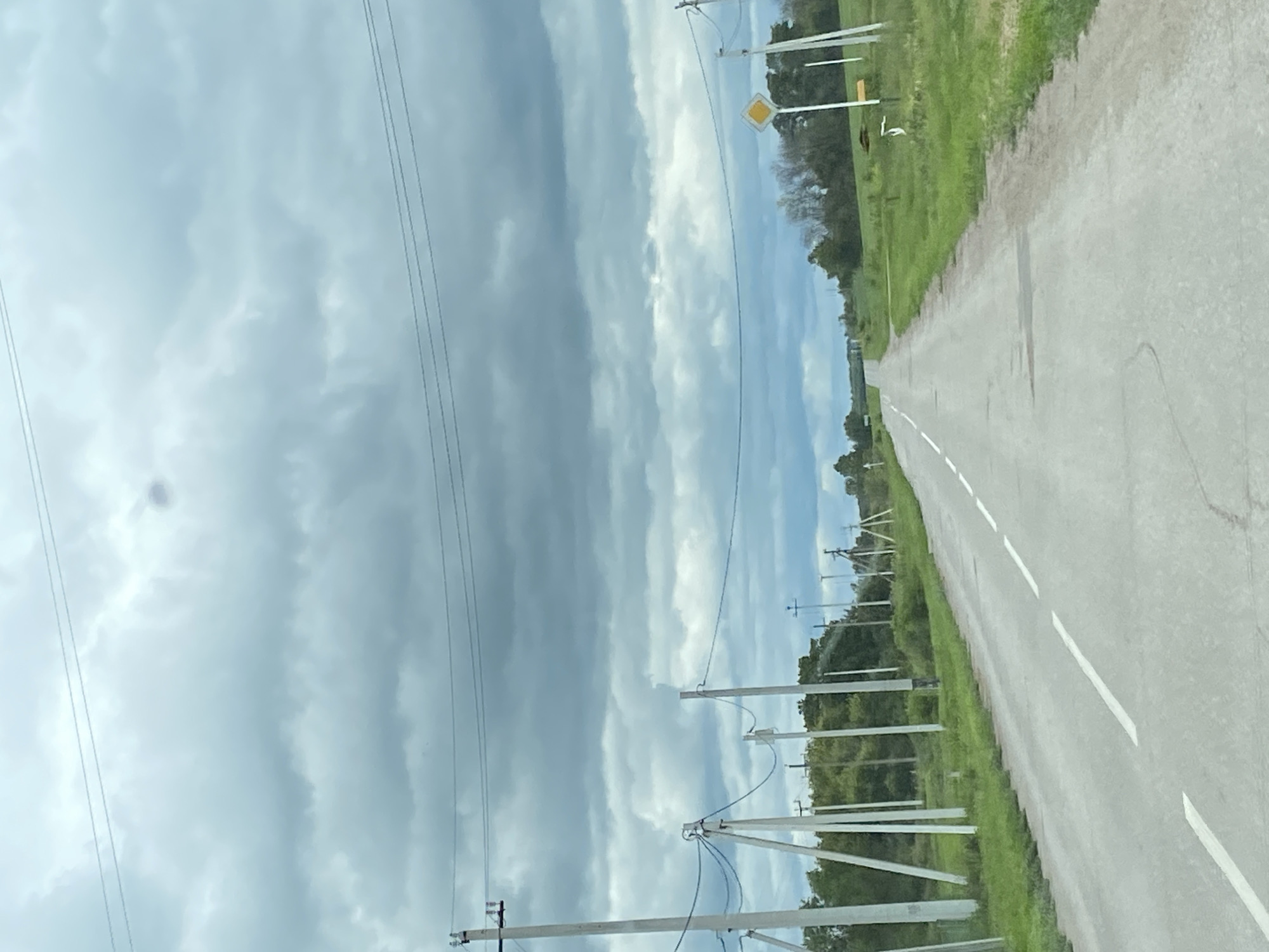 The image shows a rural road in Nadezhdino, Russia, under a cloudy sky. The road is paved and has white dashed lines marking the lanes. On either side of the road, there are grassy fields with scattered trees and utility poles. A yellow diamond-shaped road sign is visible on the right side of the road. The scene appears to be deserted, with no people or vehicles visible. The overall atmosphere is overcast and quiet.