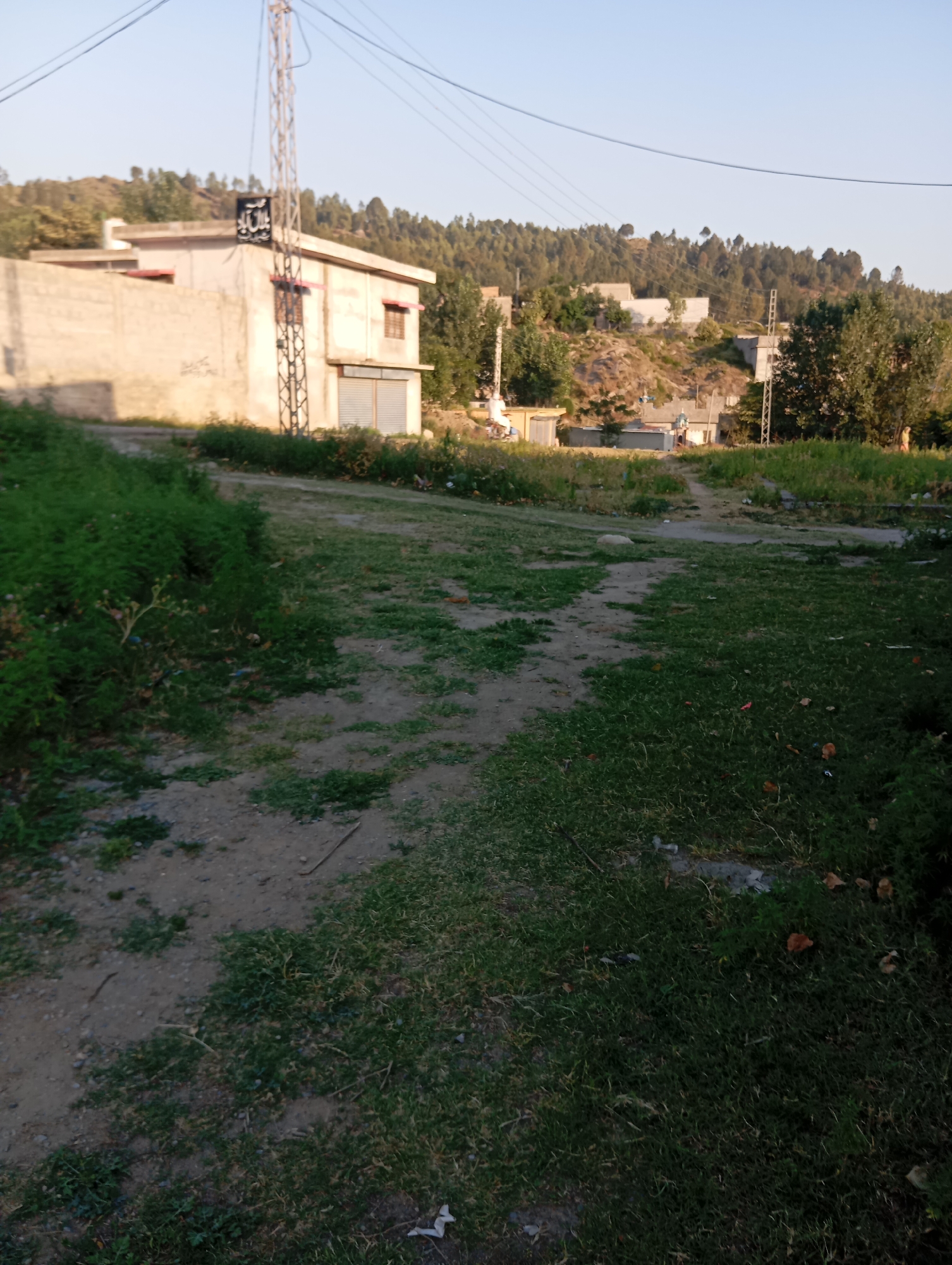 An exterior, eye-level, medium shot captures a rural landscape in Unknown, Unknown, bathed in the warm light of either sunrise or sunset. The foreground is dominated by a dirt path cutting through overgrown grass and vegetation, leading towards the midground where a light-colored, rectangular building with a light-red trim is situated. A tall metal tower with multiple crossbars stands next to the building, supporting wires that extend across the top of the frame. The building features a gated entrance and several windows, one of which is a small, square, brown-framed window. Behind the building, a stone wall, partially covered in graffiti, extends to the left. The background reveals a hillside densely populated with trees, with several other smaller buildings and structures scattered amongst them. The sky above is clear and light blue. The scene is quiet and still, with no people or animals visible.
