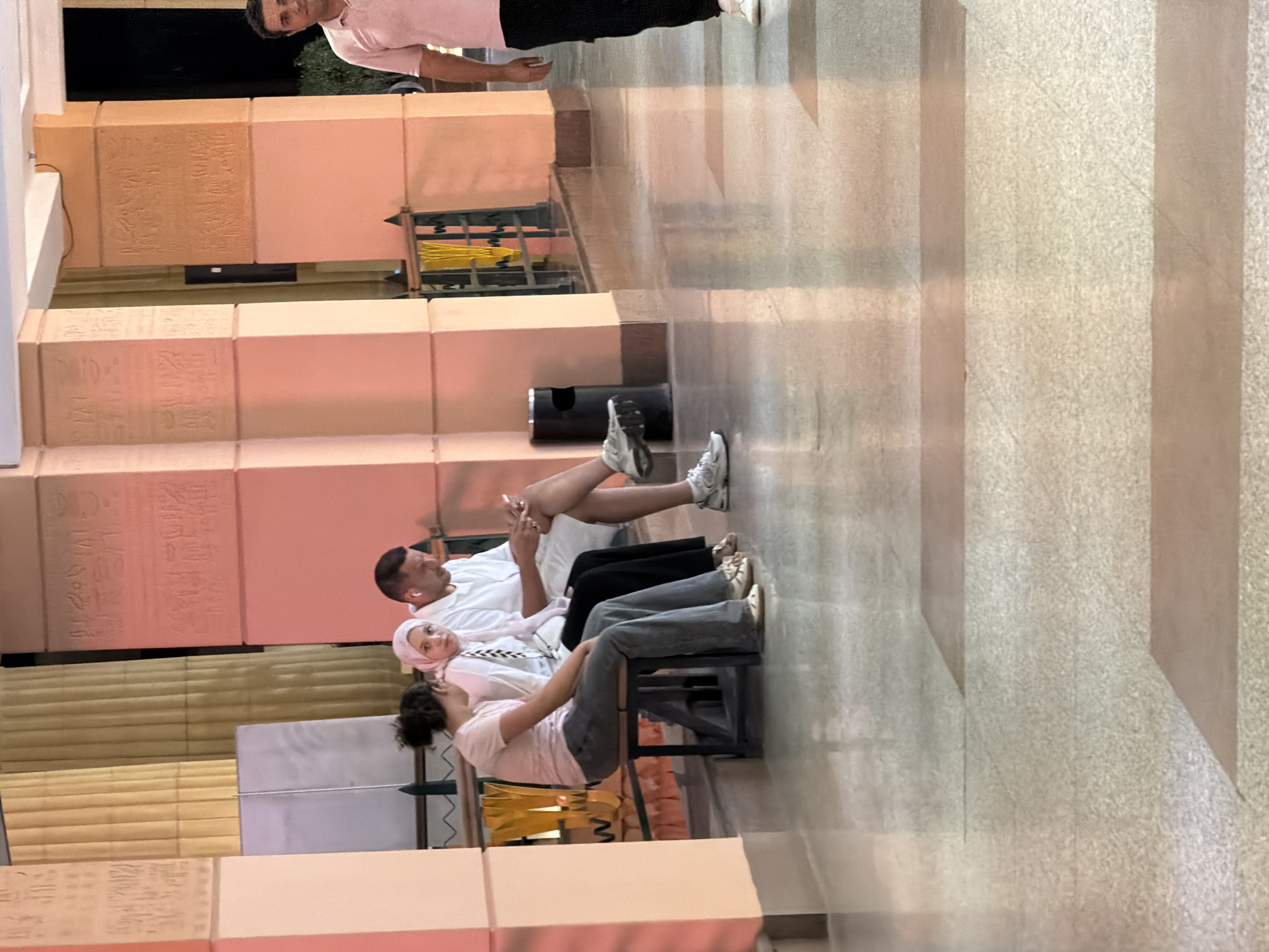 In Sharm El Sheikh, Egypt, three individuals are seated on a bench in an indoor setting with large columns decorated with hieroglyphics. On the left, a young woman with curly dark hair and a light pink t-shirt and grey pants sits facing left. Next to her, a young woman wearing a white headscarf and a white top is seated. To the right of the women, a man in a white short-sleeved shirt and dark shorts is seated, looking at a mobile phone in his hand. In the background, a man in a light pink polo shirt and black pants is walking away from the camera. The environment appears to be a hotel lobby or a similar public space, with polished floors and ornate architectural elements.