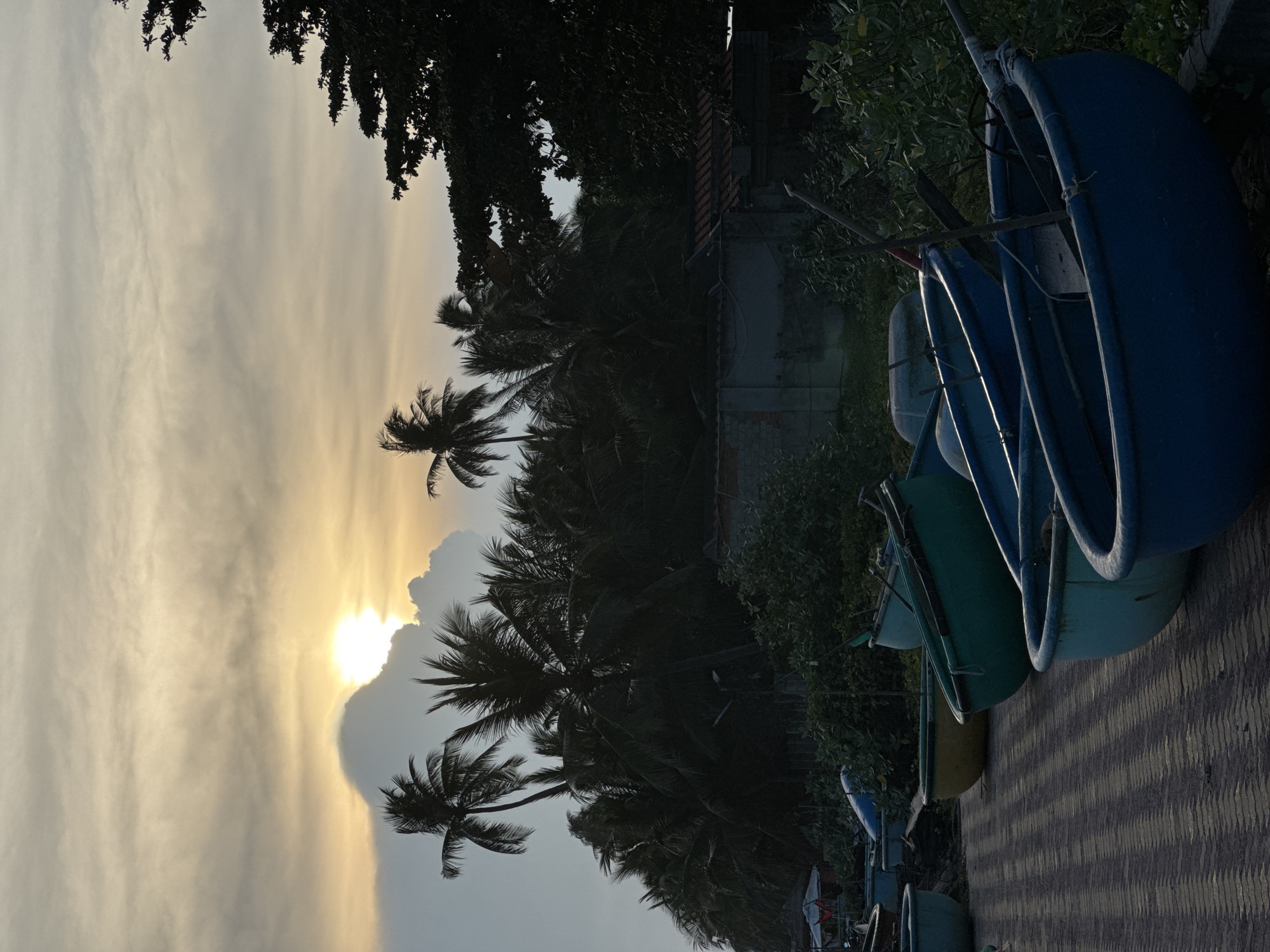 The image depicts a coastal scene in Mui Ne, Vietnam, during what appears to be either sunrise or sunset. The sky is filled with diffused sunlight breaking through clouds, casting a warm glow. In the foreground, several round, basket-like boats, commonly used in Vietnam, are arranged on a brick pathway. These boats vary in color, including shades of blue, green, and yellow. Behind the boats, lush green vegetation and palm trees create a tropical backdrop. A low-walled structure and a few scattered buildings are visible in the mid-ground. The overall impression is one of tranquil beauty and a typical coastal fishing village setting.