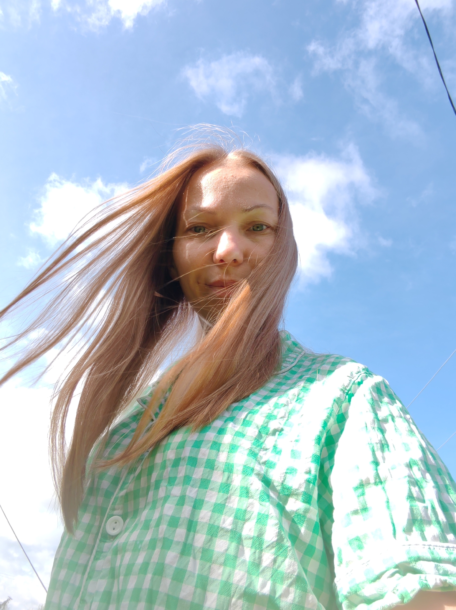 A low-angle, waist-up shot shows a woman with long, light brown hair blowing in the wind against a bright blue sky with scattered clouds. She is looking towards the camera with a neutral expression, her face partially illuminated by sunlight. She is wearing a green and white checkered shirt. The image appears to be taken outdoors, possibly in the city of Chukhloma, Russia, though no specific landmarks are visible to confirm the location. The background is dominated by the sky, with a thin black wire visible running diagonally across the upper right portion of the frame.