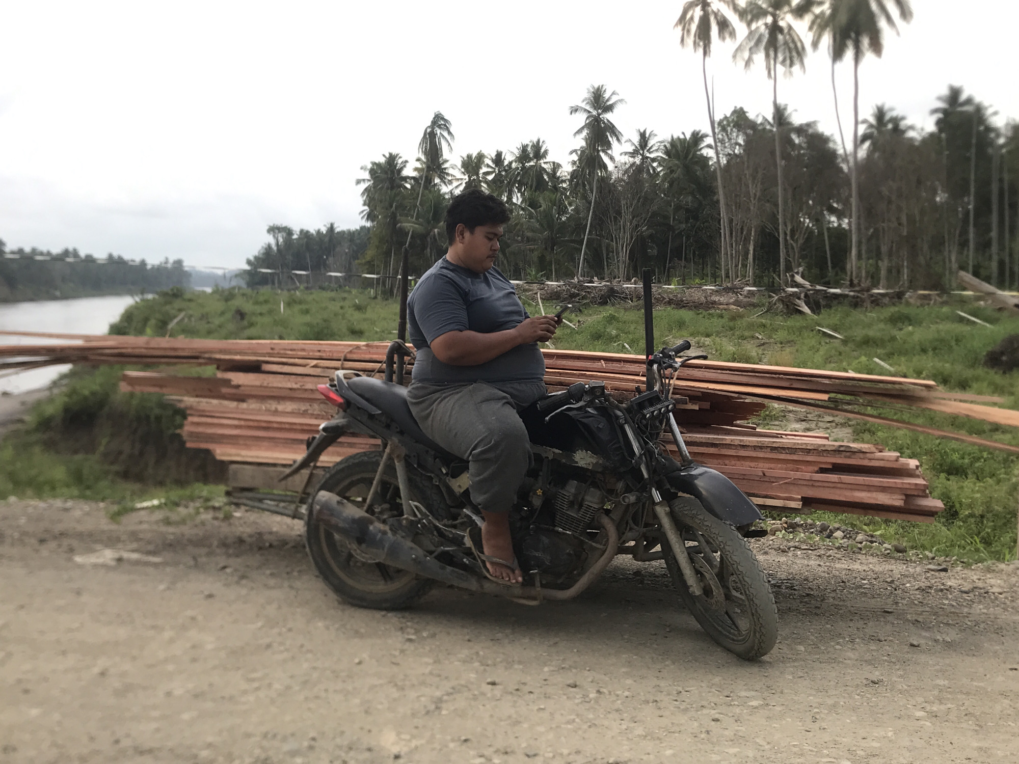 The photograph, likely taken in or near the city of Teupinreudeup, Indonesia, depicts a man seated on a weathered motorcycle. The man, wearing a gray shirt and gray pants, is looking at his phone. The motorcycle is parked on a dirt road next to stacked lumber. In the background, a river is visible, along with vegetation and palm trees. The sky is overcast.