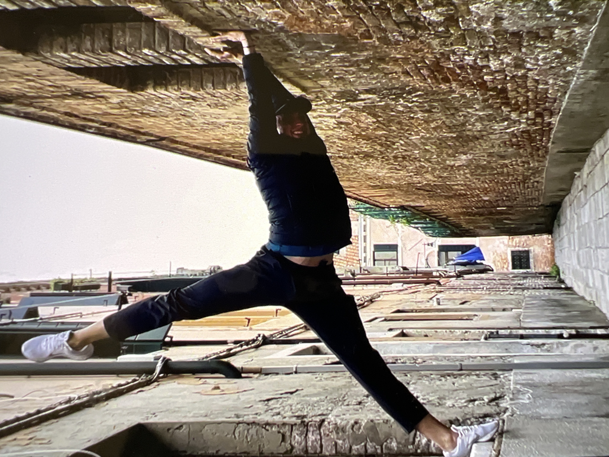 The image captures a person performing an acrobatic pose in a narrow Venetian alleyway. The person is positioned horizontally, with their legs spread wide, their left foot propped on a ledge against a textured wall and their right hand pressed against a brick wall. They are wearing dark clothing and white sneakers. The alley is lined with old buildings, one with a weathered stone facade and the other with a rough brick exterior. The sky is overcast. The scene is set in Venice, Italy.