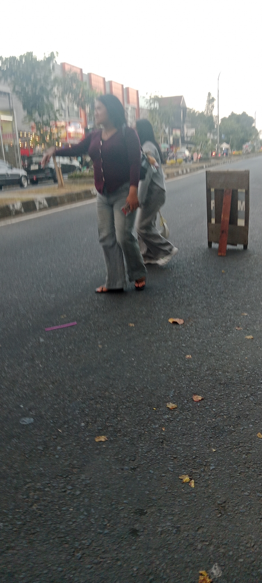 In the city of Ngemplak, Indonesia, two women stand on an asphalt road. The woman in the foreground, on the left, wears a maroon cardigan and light-colored flared pants. She is facing to her right, with her left arm extended as if hailing a vehicle or gesturing. Her right hand holds a dark object, possibly a phone. She is wearing sandals. Behind her, a second woman, also wearing light-colored pants, is seen from the back, walking away from the camera. She carries a bag on her left shoulder. To the right of the women, a wooden traffic barrier with red and white markings stands on the road. In the background, buildings with orange facades are visible, along with trees and streetlights. The sky is overcast. Leaves are scattered on the road surface.