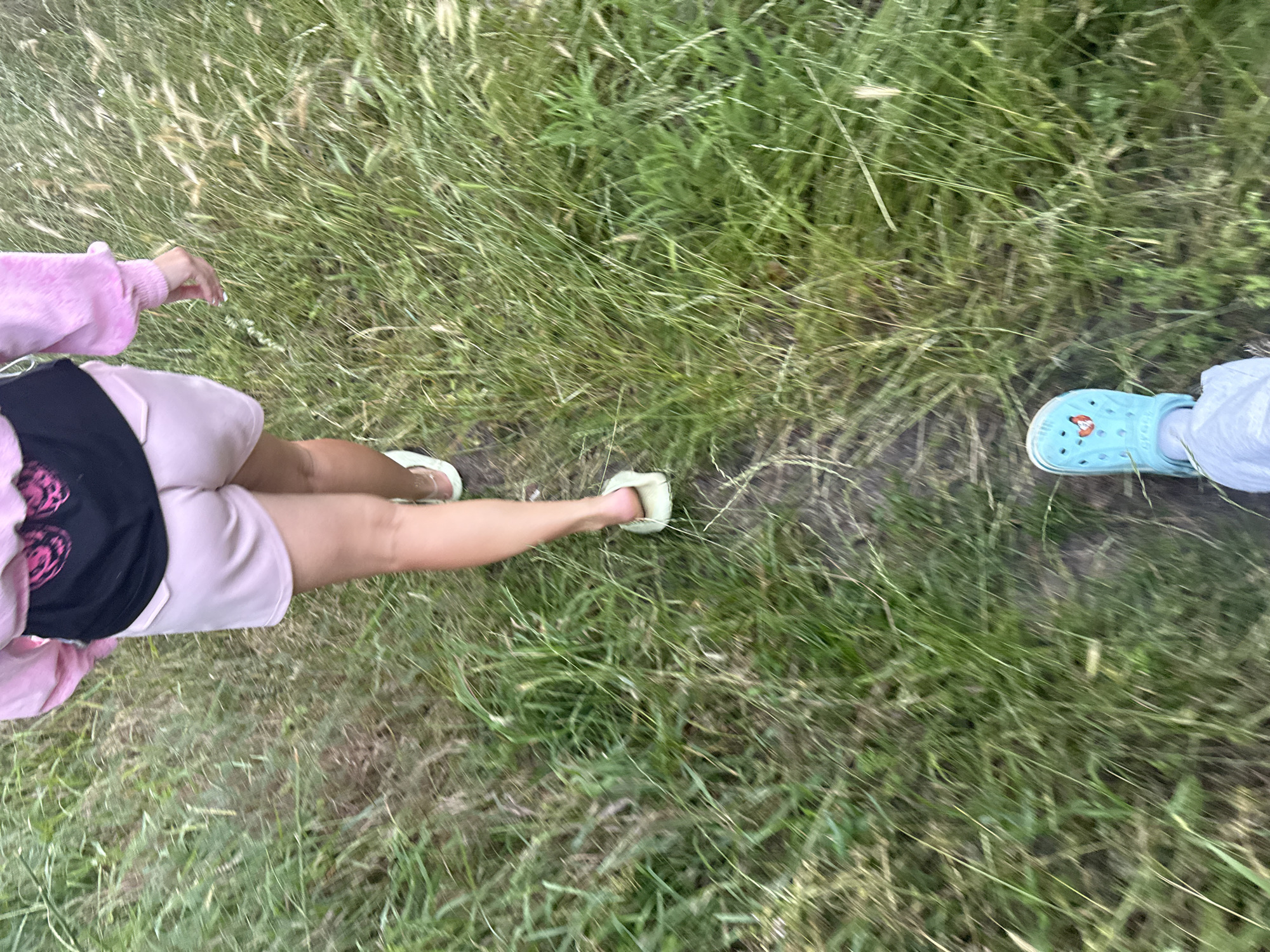 The image depicts a ground-level perspective of two people walking on a dirt path through tall green grass. The person in the foreground is wearing light blue Crocs with a cartoon character graphic. The person further ahead is wearing pale pink shorts, a black t-shirt with a pink graphic on the back, and a pink fuzzy jacket. They are also wearing light green slip-on shoes. The scene is outdoors, with soft natural lighting and a slightly blurred effect suggesting movement. The environment is natural and appears to be a field or meadow. The specific location within the city of Obukhivka, Ukraine, cannot be determined from the visual information provided.