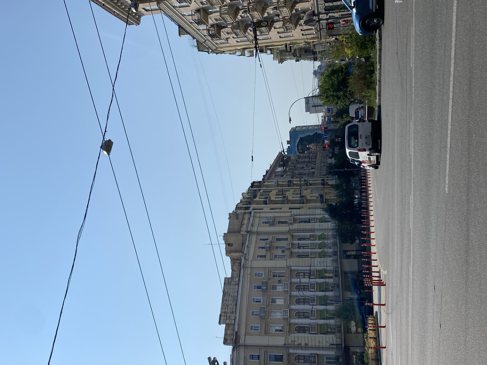 The image shows a wide street in a city with ornate, multi-story buildings lining both sides. The buildings have a classical architectural style with arched windows and balconies. Overhead power lines crisscross the sky, which is clear and blue. Traffic cones are placed in a row along the side of the road, and several cars are visible, including a white van and a blue SUV. A traffic light is also visible. The scene appears to be a daytime shot in an urban environment.