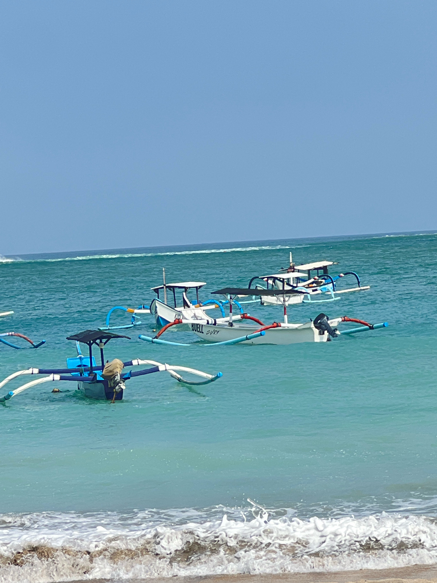 In Kuta, Indonesia, several traditional outrigger boats are moored on the turquoise waters of the ocean. The boats, with their distinctive outrigger pontoons, are painted in various colors, including white, blue, and accents of red and yellow. One boat in the foreground has a blue hull and a black canopy, with a tan bag secured to its stern. Another boat, further out, is white with a blue and white canopy. The water is clear, showing a gentle wave breaking on the sandy shore in the foreground. The sky above is a clear, pale blue.