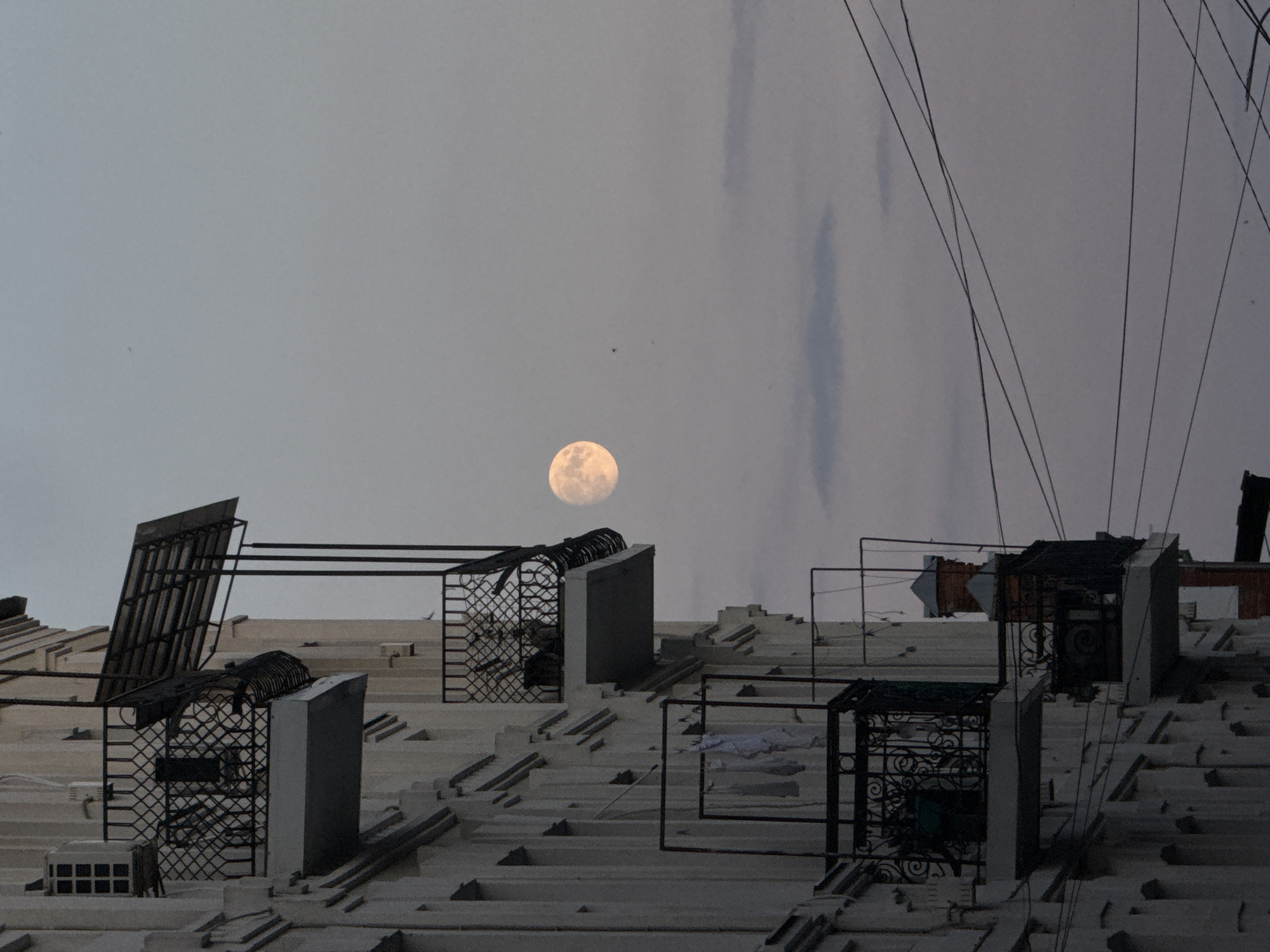 The image shows a residential building with several balconies, one of which is draped with laundry. The full moon is visible in the dusk sky, partially obscured by a balcony awning. The scene is likely from Vinnytsia, Ukraine, based on the architectural style and the presence of overhead wires. The overall atmosphere is calm and serene, with the soft light of the moon casting a gentle glow on the building. The image is composed from a low angle, emphasizing the height of the building and the vastness of the sky. There are no people visible in the image. The image appears to be a photograph taken during the twilight hours.