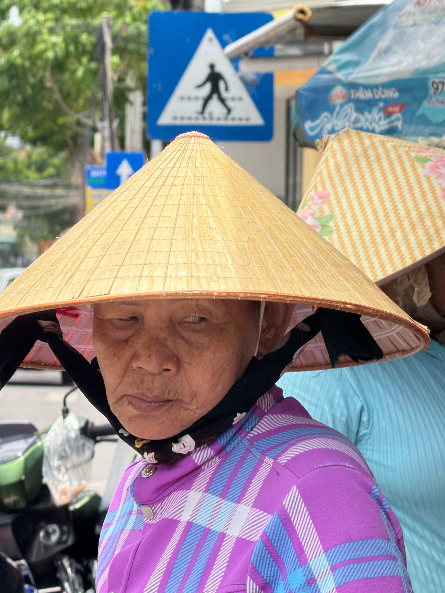 A close-up, eye-level shot shows an elderly Vietnamese woman wearing a conical hat and a plaid shirt in Nha Trang, Vietnam. The woman's face is visible beneath the brim of her hat, with wrinkled skin and a neutral expression. She is wearing a purple and blue plaid shirt and a black scarf around her neck. In the background, a blue pedestrian crossing sign with a silhouette of a person walking is visible. Another conical hat, adorned with a floral pattern, is partially visible to the right of the main subject. The scene appears to be outdoors, possibly on a street, given the presence of traffic signs and the background blur suggesting movement.