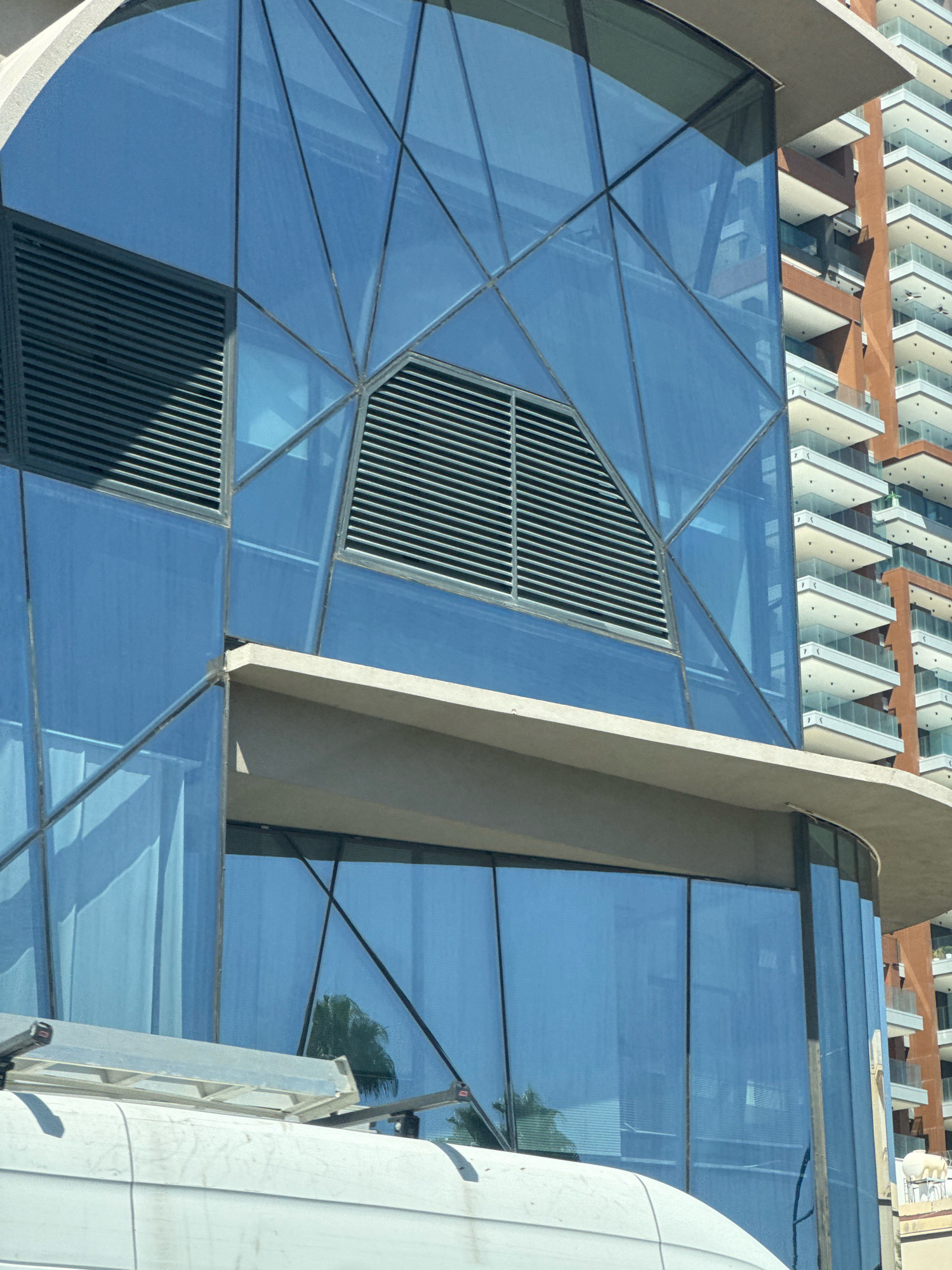 The image captures a close-up view of a modern building with a striking geometric facade constructed of blue reflective glass. Thin, dark metal framing divides the glass panels into a complex pattern. Two large rectangular vents with horizontal louvers are integrated into the facade, one positioned higher and slightly to the left, the other lower and more centrally. Above and to the right of the main building, the upper floors of a taller, older building are visible. This adjacent structure has a more traditional design, featuring rows of balconies with white railings and an orange-brown brick exterior. A light-colored, curved architectural element, possibly a balcony or awning, juts out from the blue glass building, creating a horizontal division. In the foreground, the top of a white vehicle, likely a van or truck, is visible. A metal ladder or rack is mounted on top of this vehicle. Reflections of palm trees can be seen in the blue glass of the modern building, hinting at the surrounding environment. The scene is set in the city of Agios Tychona Community, Cyprus.